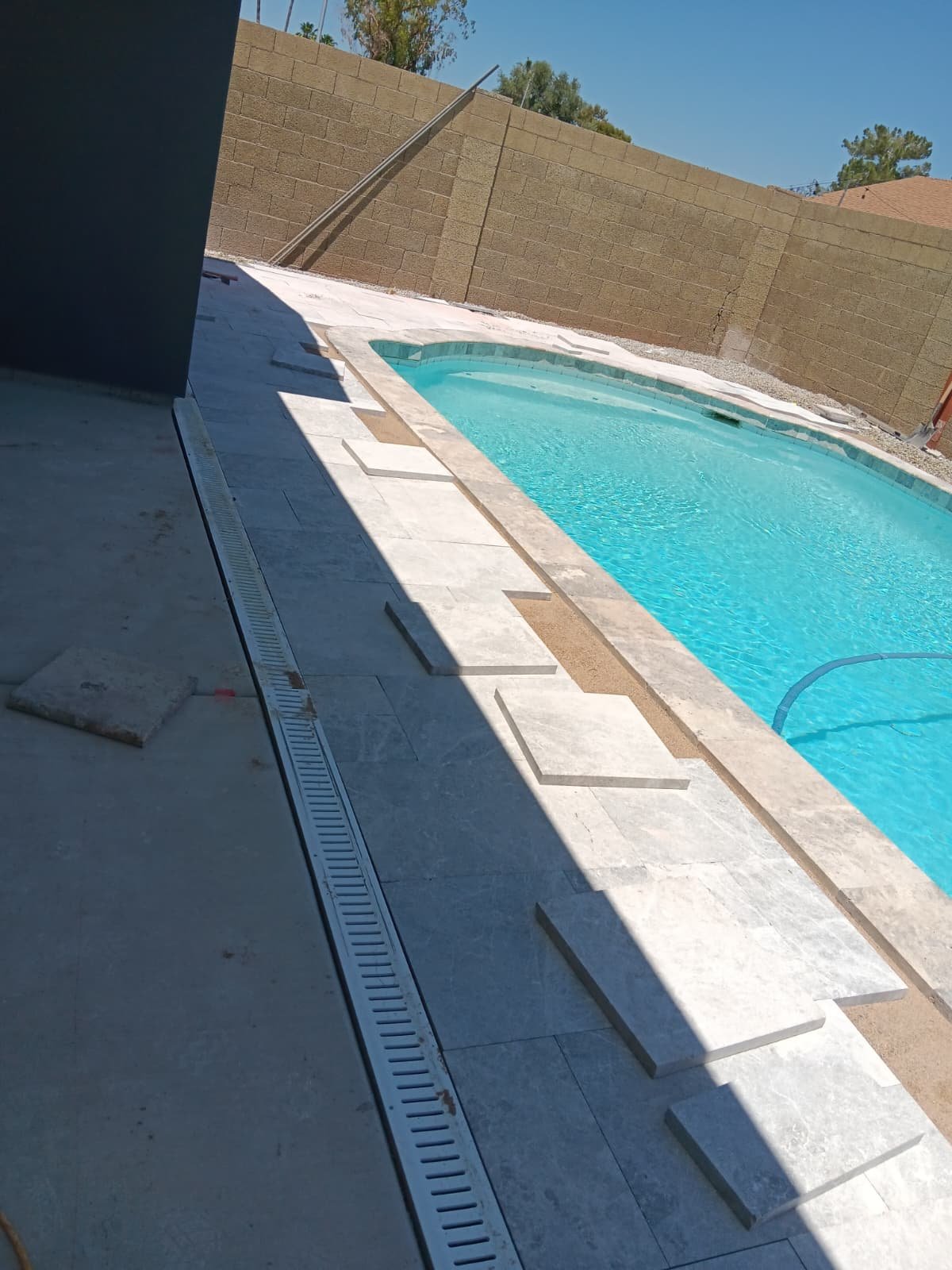A backyard with a swimming pool, partially covered by shadows, with a brown brick wall surrounding the area and a cloudy sky above.