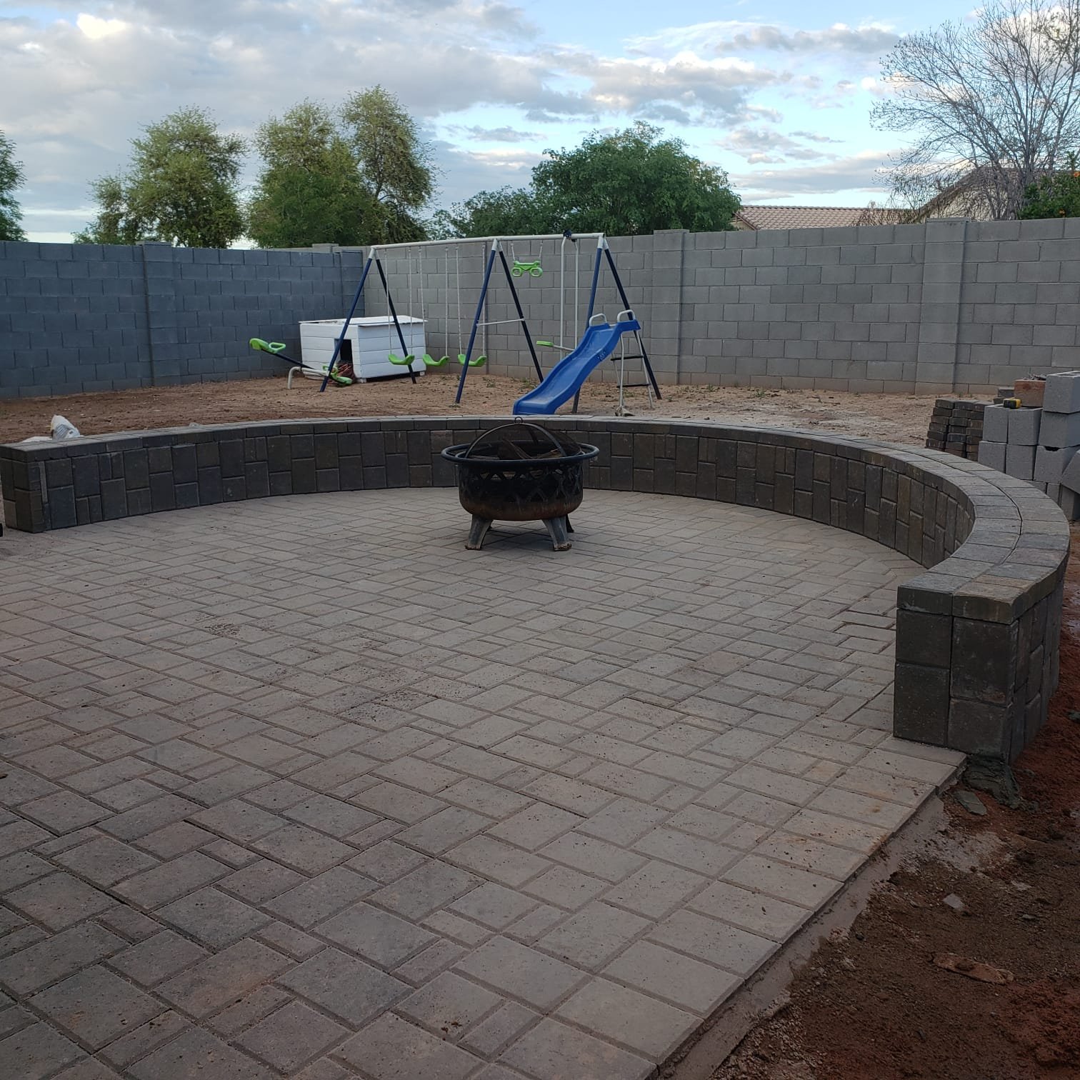 Backyard with a paved patio, a fire pit in the center, a raised brick border, and a gravel area with a children's swing set and slide against a cinder block fence, with trees and a cloudy sky in the background.