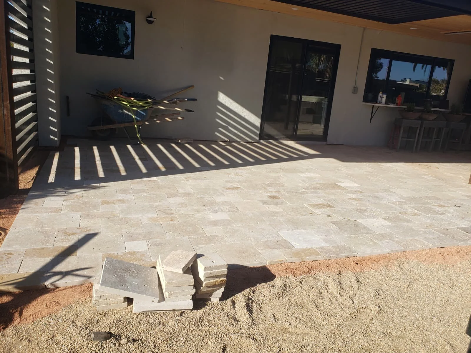A newly constructed outdoor patio with light-colored stone tiles, with some construction materials in the foreground, and a small wheelbarrow with tools and hoses against a wall near a sliding glass door and window.