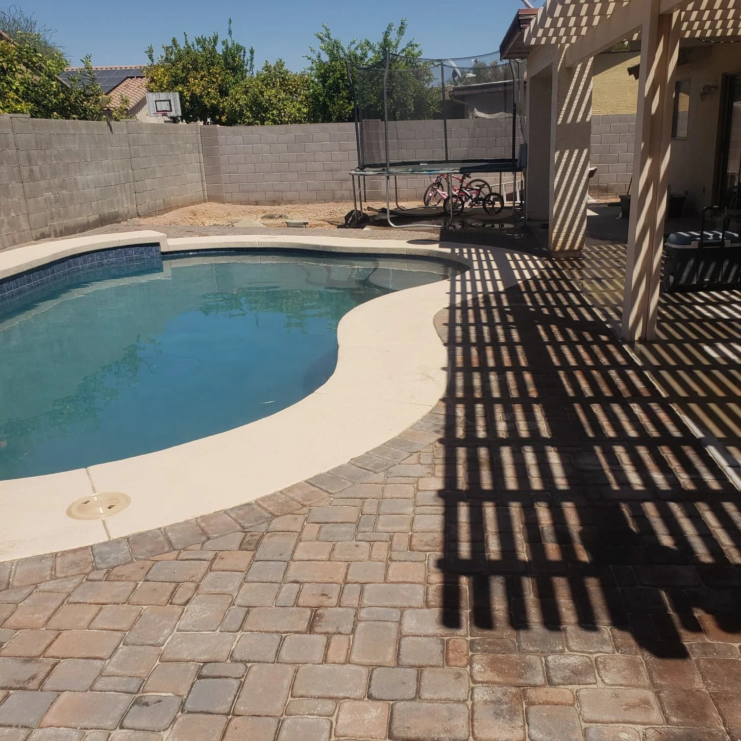 Backyard with a kidney-shaped swimming pool, brick paving, a shaded patio area with lattice shadows, a trampoline, bicycles, and a cinder block wall with trees in the background.