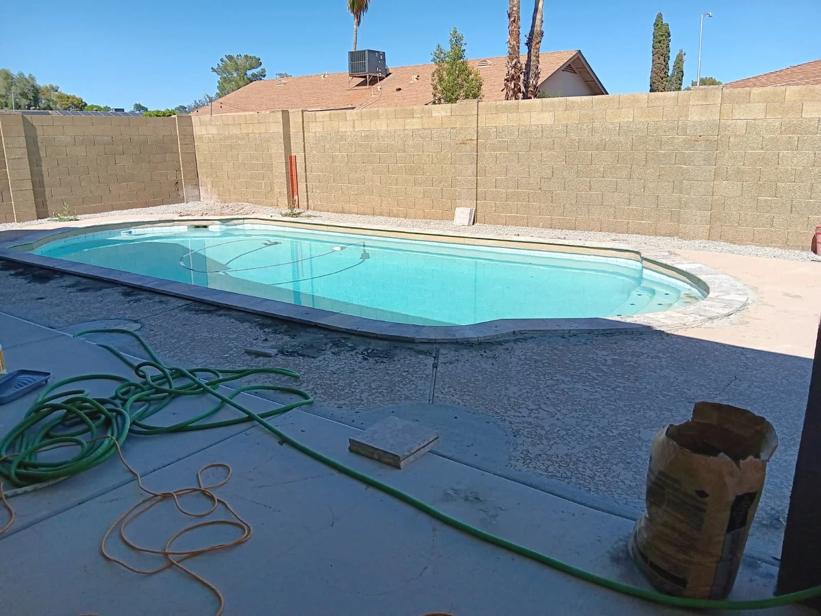 Residential backyard with a side view of an in-ground swimming pool, surrounded by a concrete deck, with a brick wall fence, some trees, and neighboring houses in the background. Pool cleaning tools and hose are on the deck.