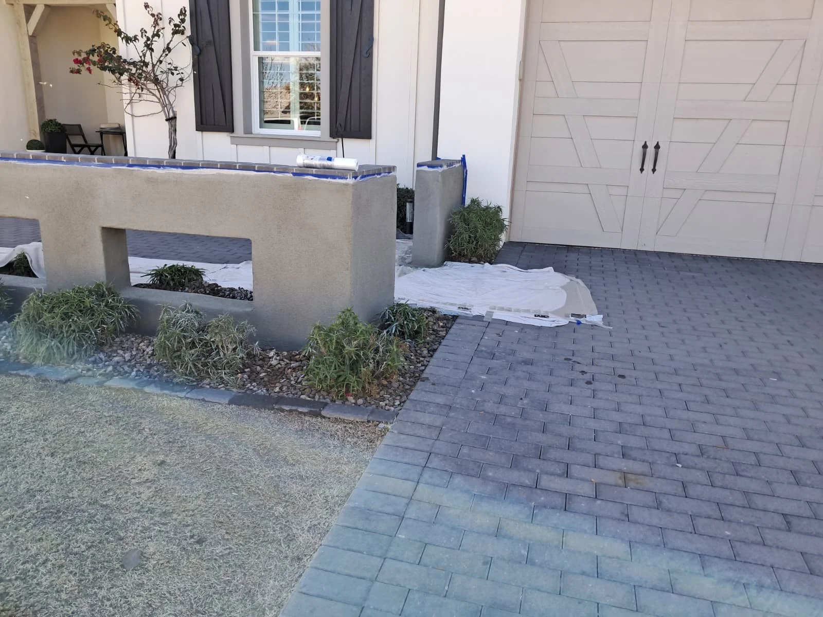 Exterior of a house under construction or renovation with potted plants, a small garden with bushes, a white wall, a window with shutters, a shed door, and construction materials scattered on the ground.