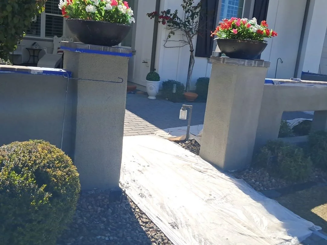 A front yard entrance with two large flower pots filled with pink, white, and red flowers, a pathway covered with white fabric or tarp, and potted plants near a house with blue shutters.