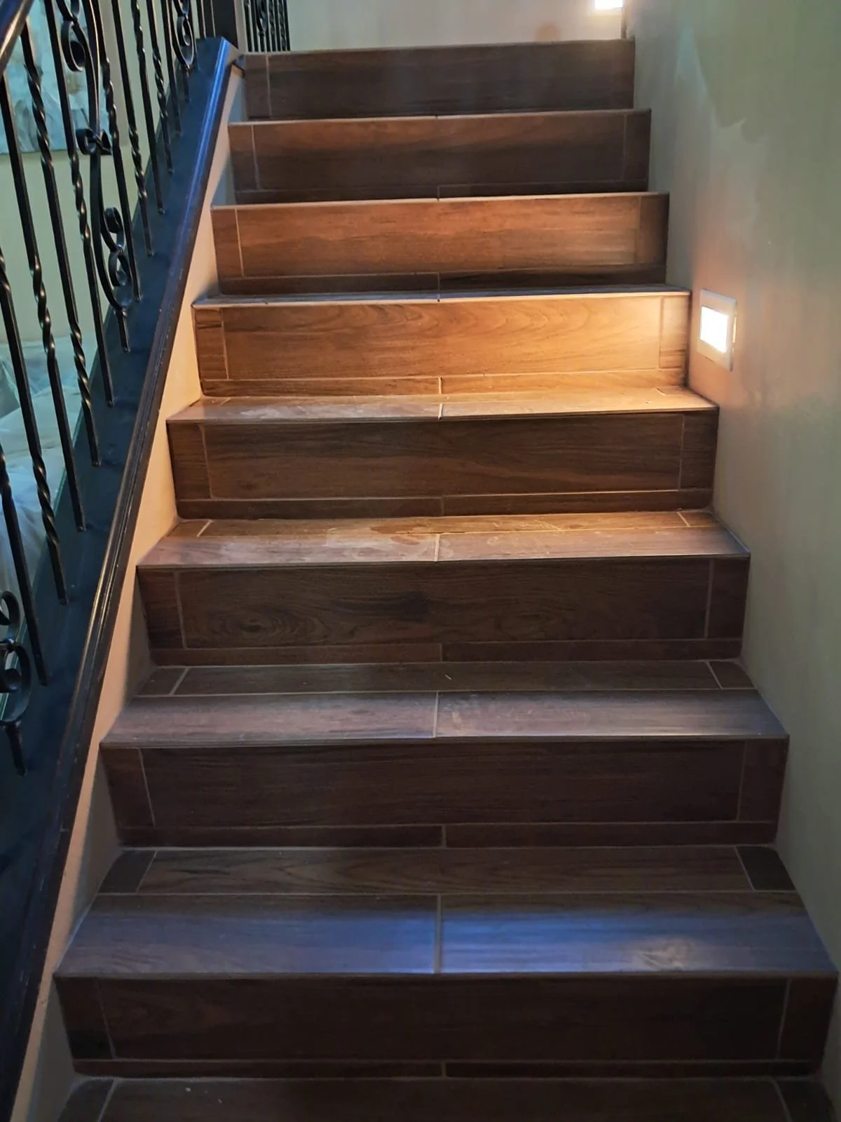Indoor staircase with dark wood steps, black metal railing on the left, green wall on the right, and a small floor light on the right wall.