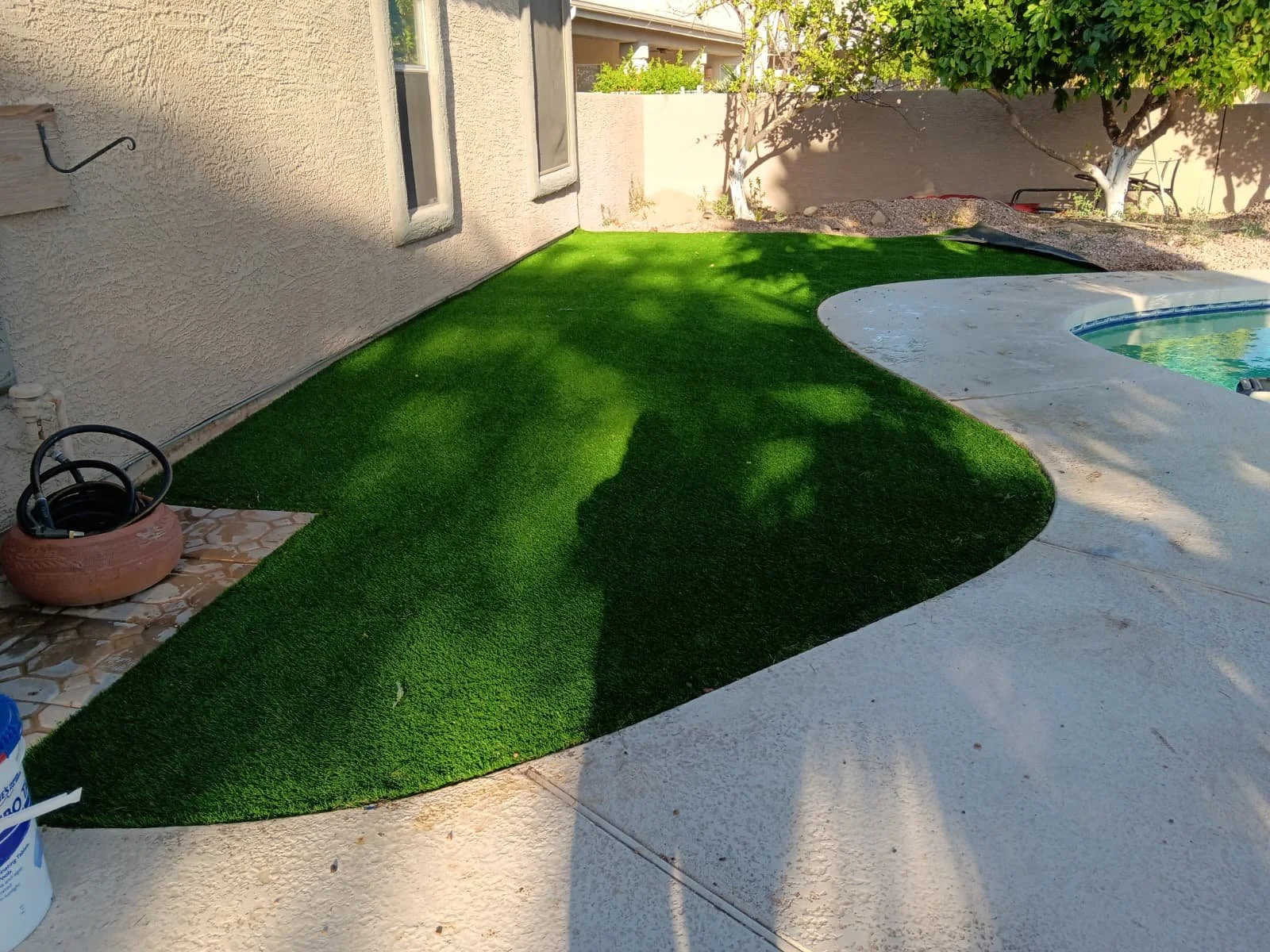 A backyard with an artificial grass patch lining a concrete patio near a swimming pool, with a house wall on the left, trees providing shade, and a bucket and garden tools in the scene.