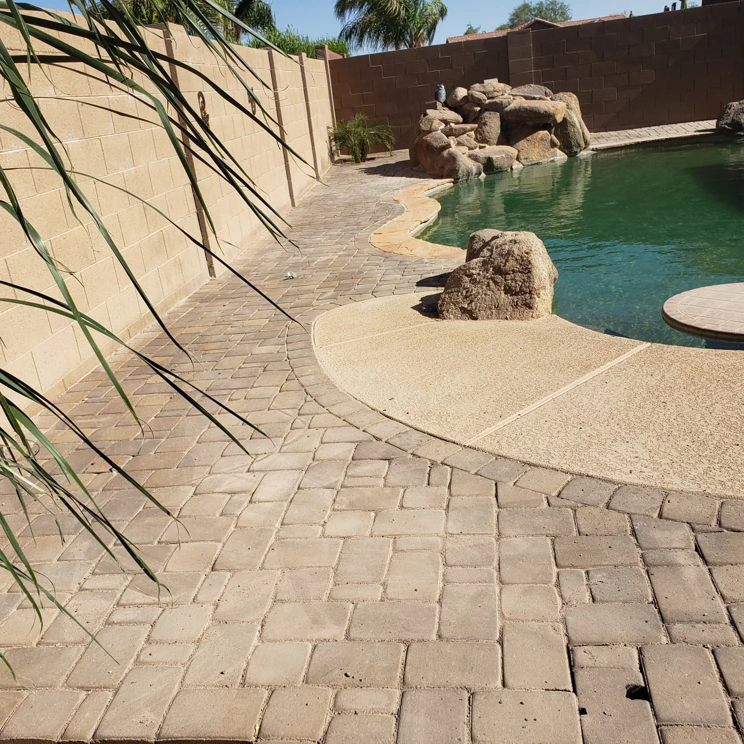 Backyard pool area with stone paver deck, large rocks, and a brick wall fence. Trees and plants are visible in the background.