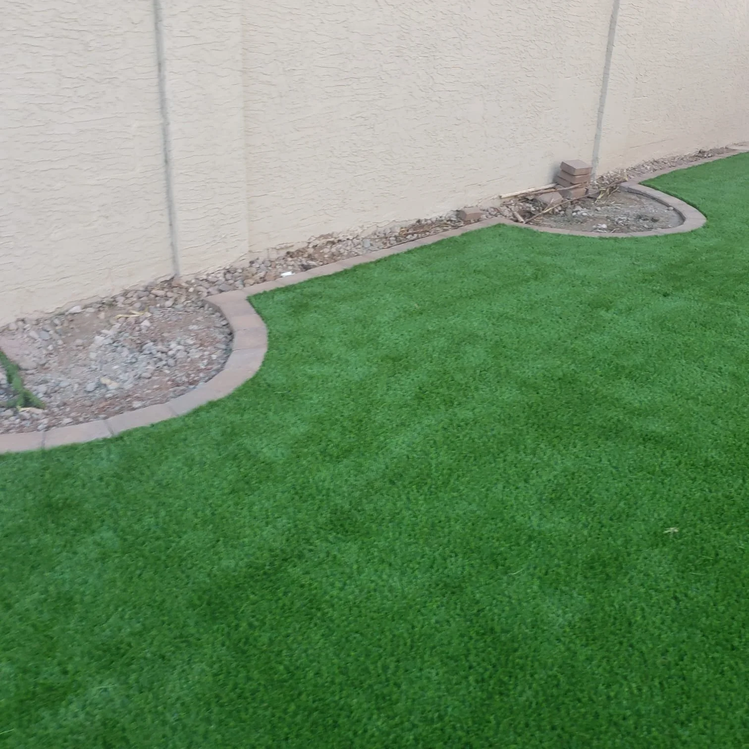 Part of a backyard with bright green artificial grass and a beige stucco wall. There are small shrub beds with stones near the wall, some bricks are stacked at the edge of the shrub beds, and a section of dirt is visible in the beds.