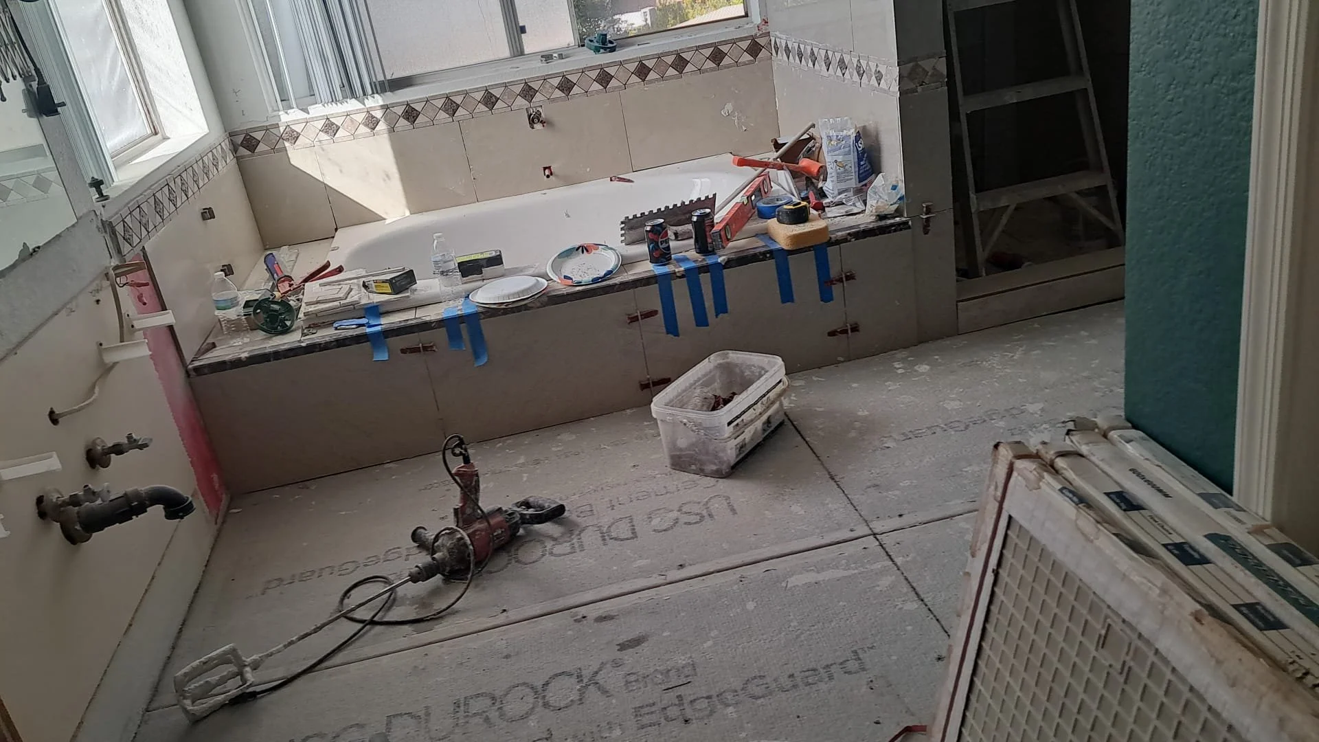 A bathroom under construction with tools and materials, including a portable heater, taped-off tiles, bare floor with insulation, and a partially installed bathtub.