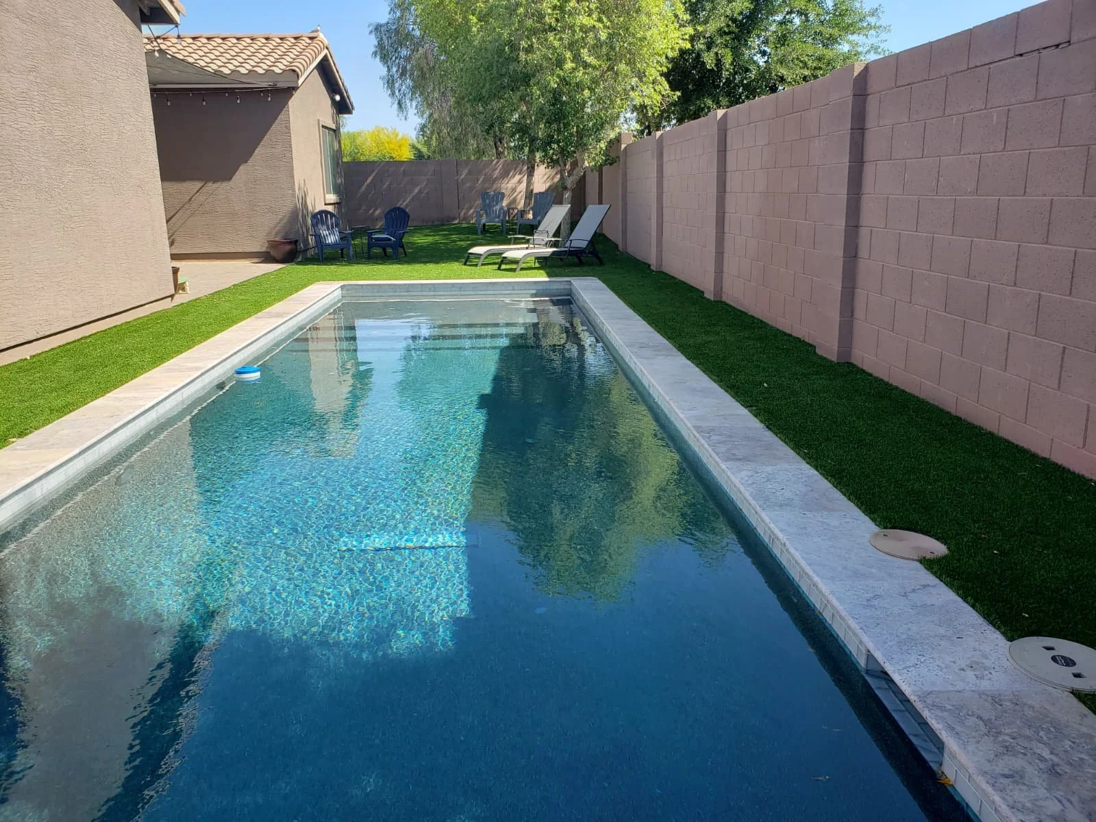 Backyard with a rectangular swimming pool, green artificial turf, and outdoor seating including poolside chairs and table, enclosed by a beige brick wall, with a house in the background and trees.