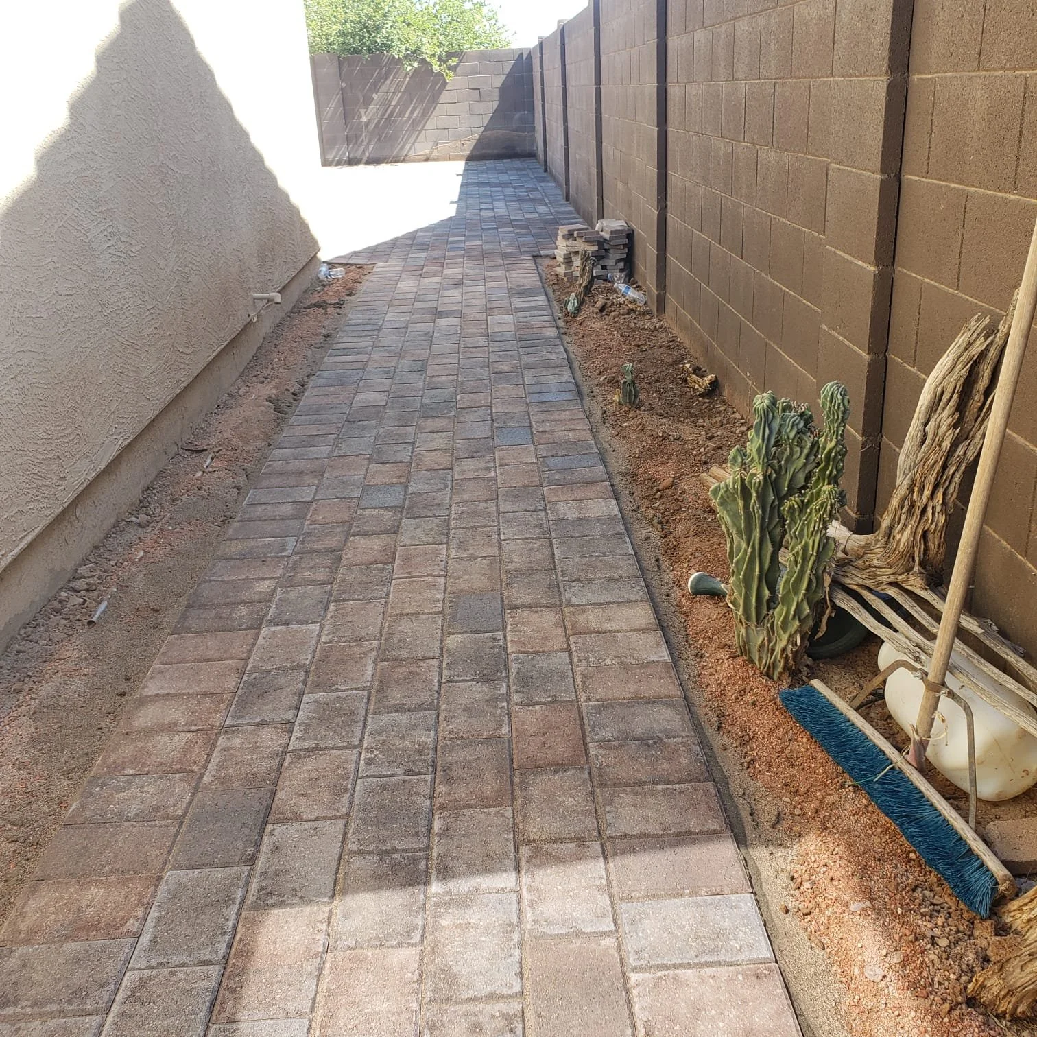 Newly paved brick sidewalk alongside a brown brick wall and a stucco wall, with soil and plants on the right side.
