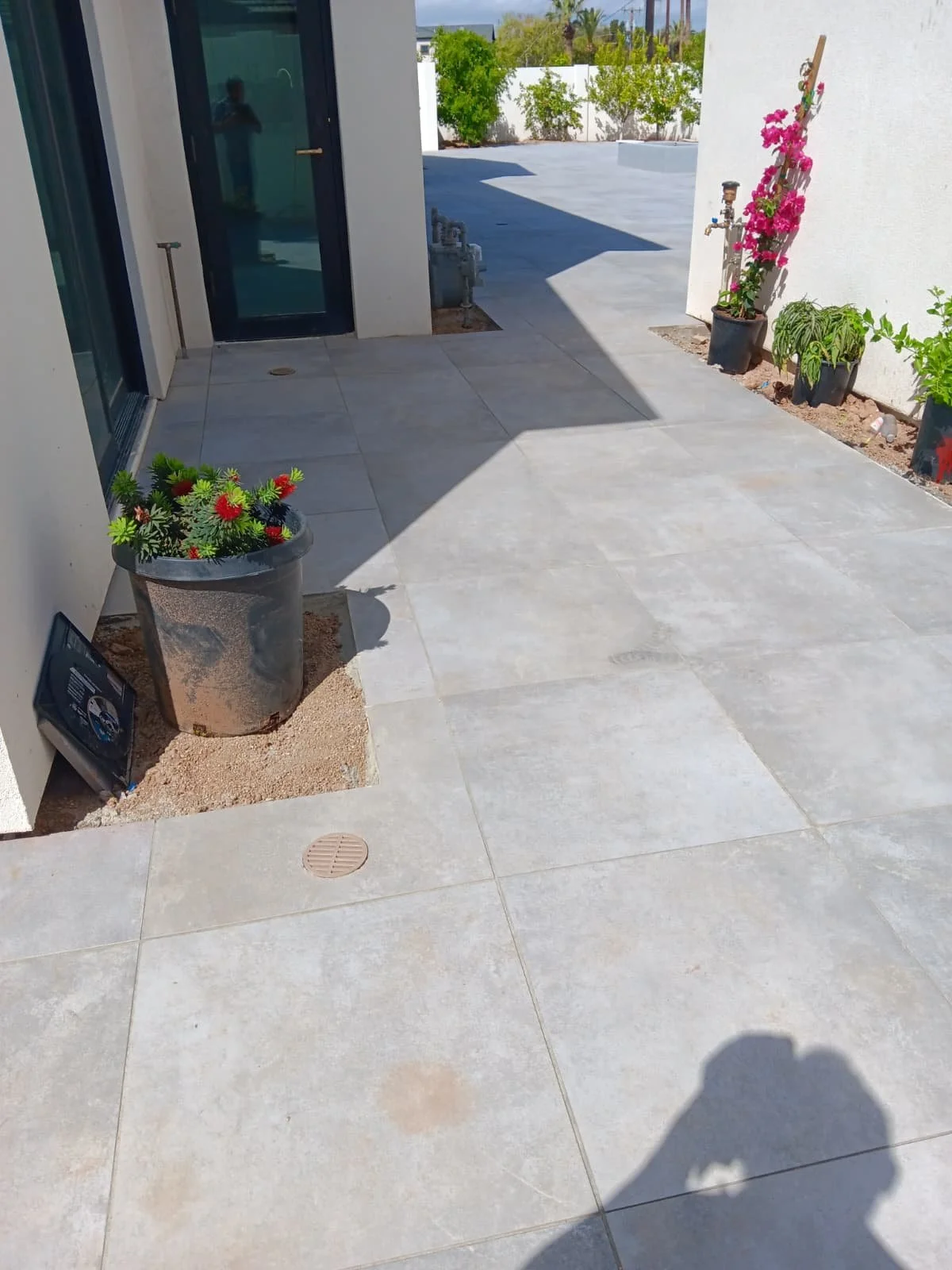 View of an outdoor patio area with large light-colored tiles, potted plants, and a white wall with a garden in the background. Sunlight casting shadows on the ground.