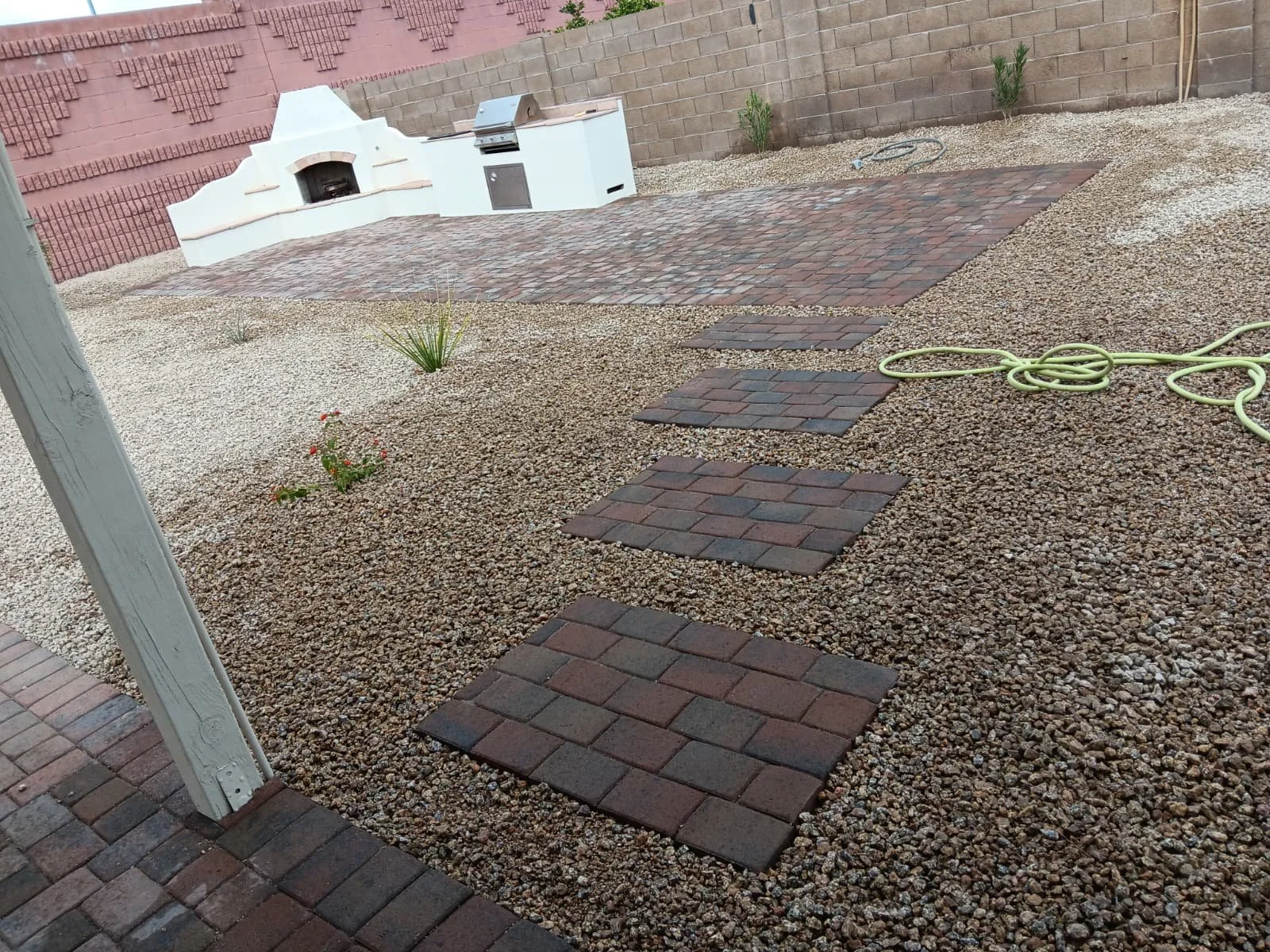 A backyard with stepping stone pavers on gravel, a small grill area with a brick surface, a garden hose, and a brick wall with some plants.