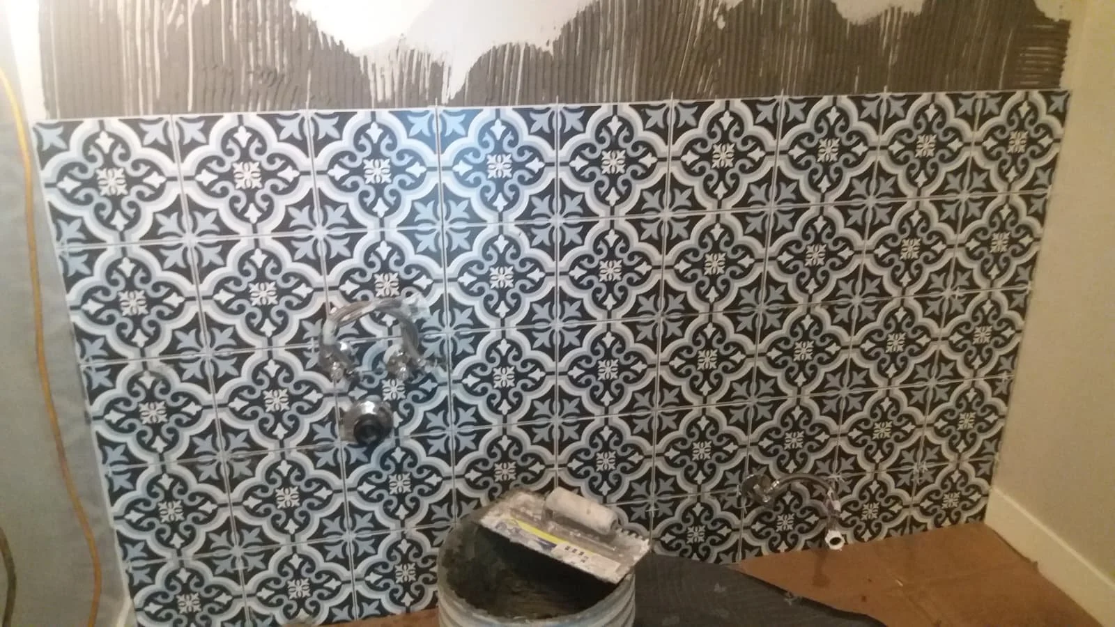 Interior wall with patterned black and white tile, partially with wooden panels, next to a bucket of tile adhesive and some tools, likely in a bathroom or kitchen renovation.