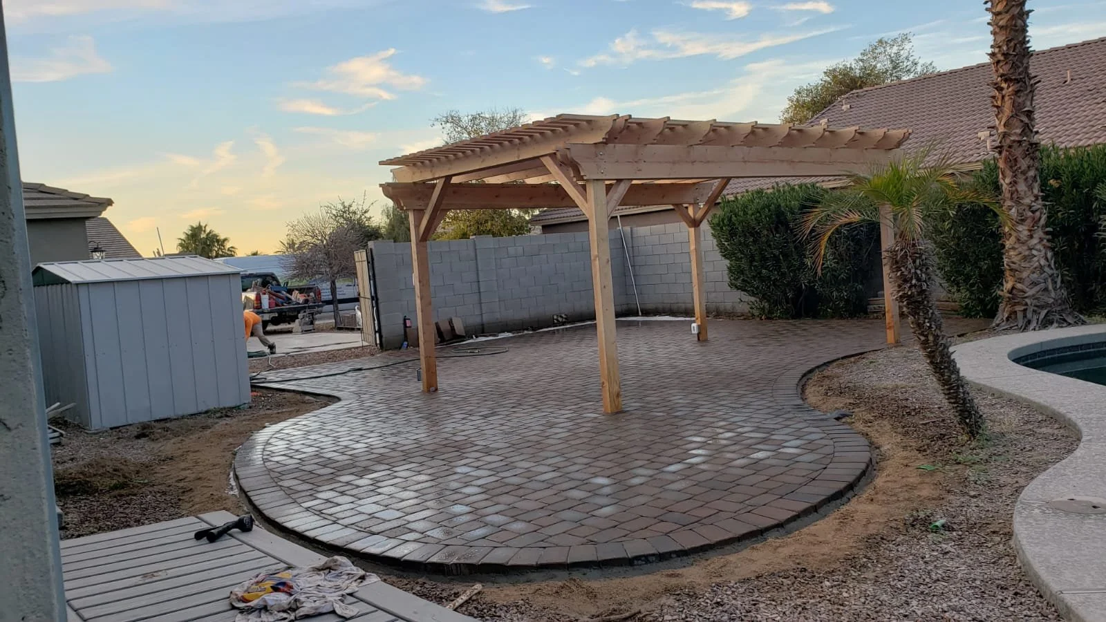 Backyard patio under construction with a new paved brick surface, a wooden pergola frame, and desert plants including palm trees, with a brick wall, a metal shed, and neighboring houses in the background.