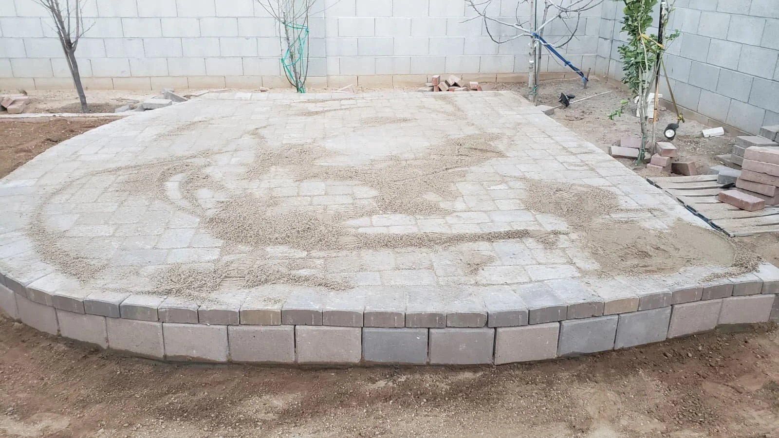 Paved patio area under construction with interlocking paving stones, some loose sand, and an unfinished border of larger pavers, surrounded by bare dirt and a brick wall in the background.