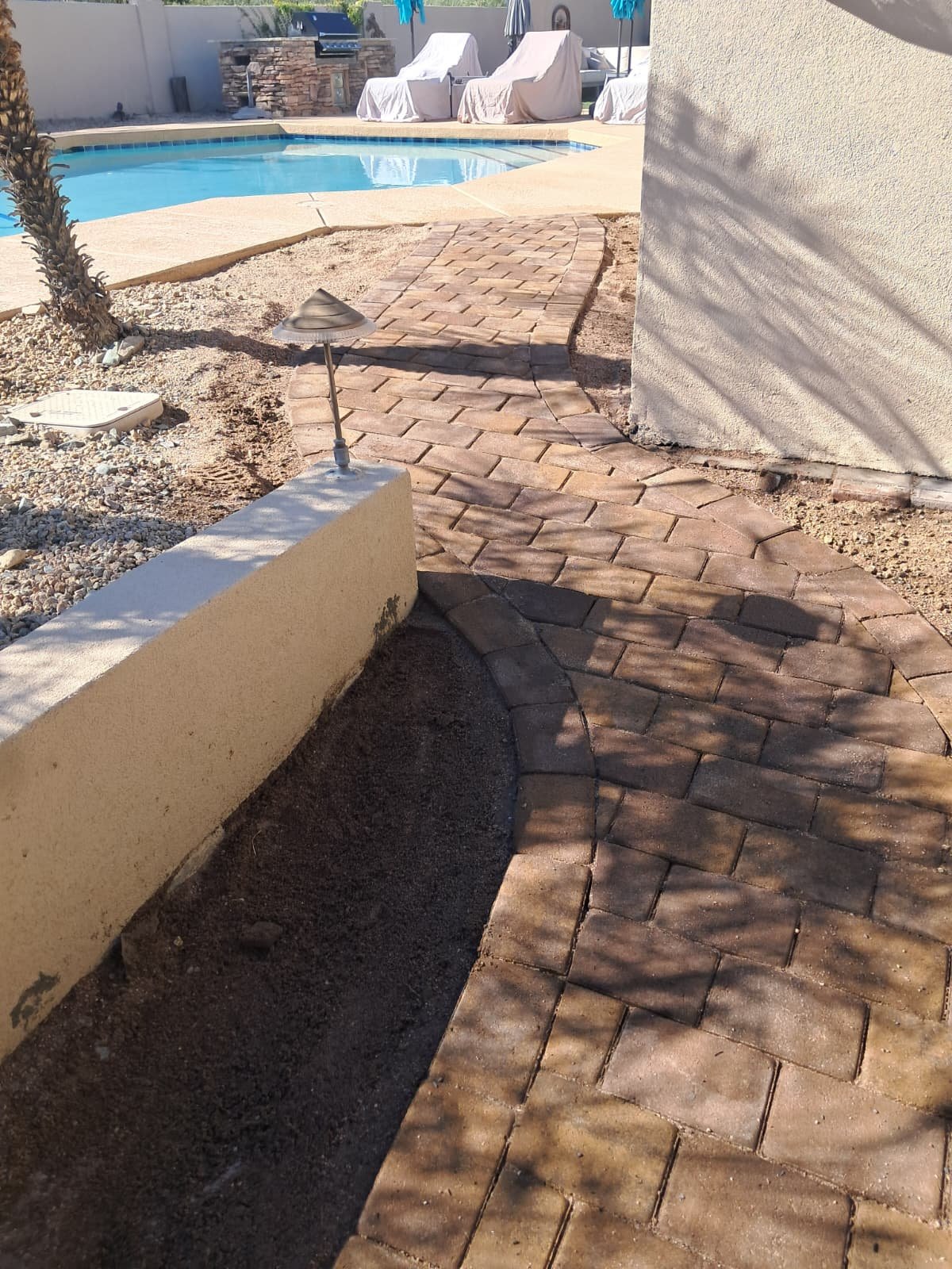A backyard scene with a curved brick pathway leading to a swimming pool. The pool is surrounded by lounge chairs covered with white towels and a stone outdoor kitchen area. There are shadows cast on the wall and ground.
