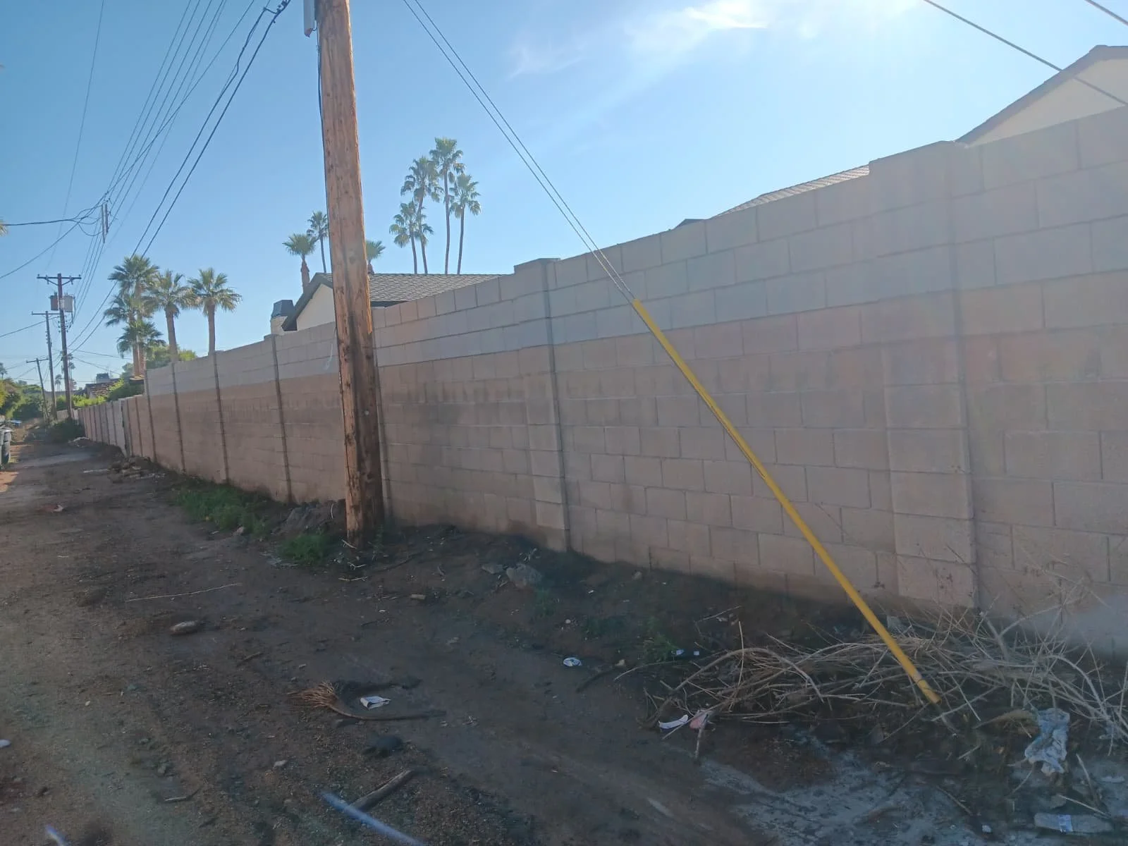 A dirt road runs alongside a tall brick wall, with utility poles and power lines stretching overhead. Palm trees are visible in the background under a clear blue sky.
