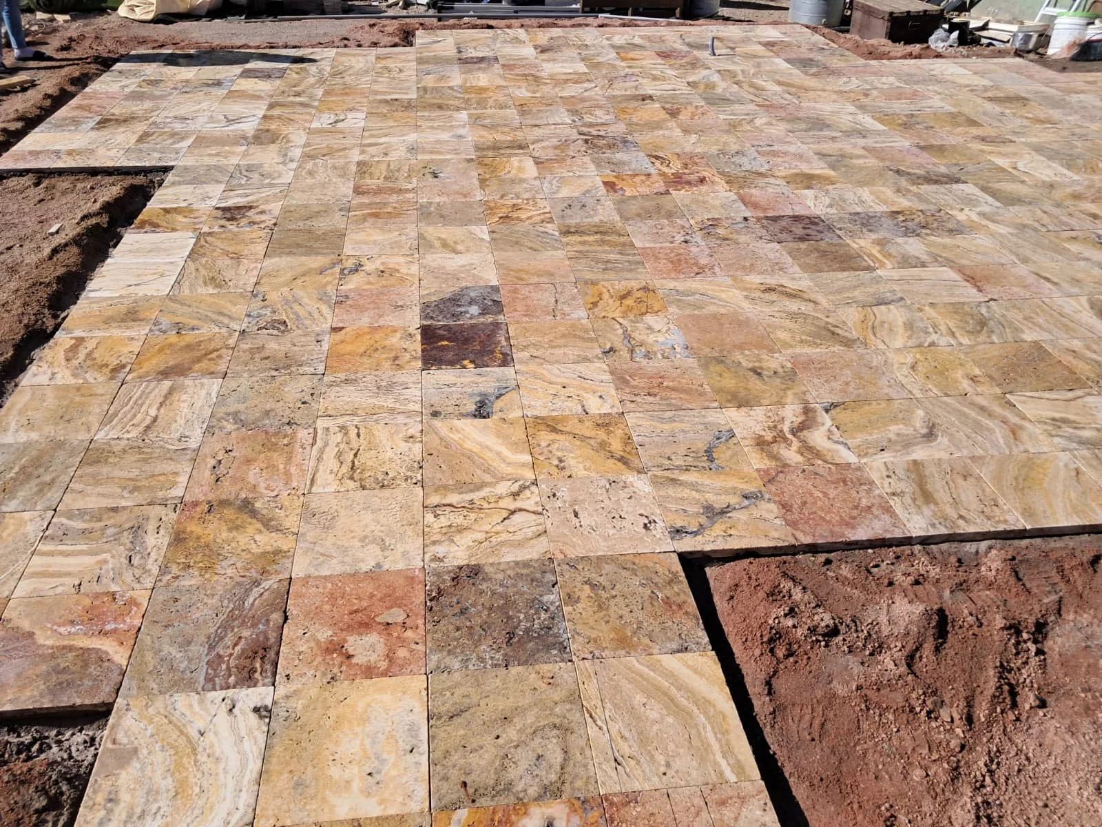 A partially completed stone patio with square tiles in natural earth tones, surrounded by exposed soil and construction materials.