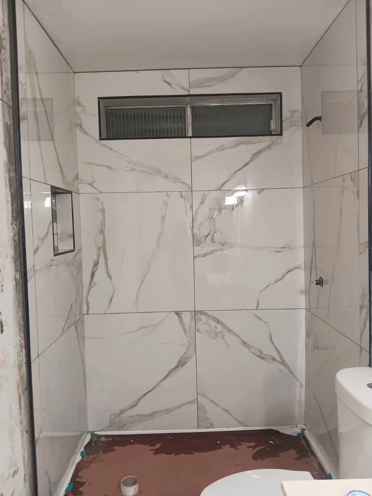 A bathroom shower area with white marble tiles with gray veining, a horizontal window near the ceiling, a small built-in shelf, and a partially visible toilet on the right. The floor is still under construction with some tools and materials.