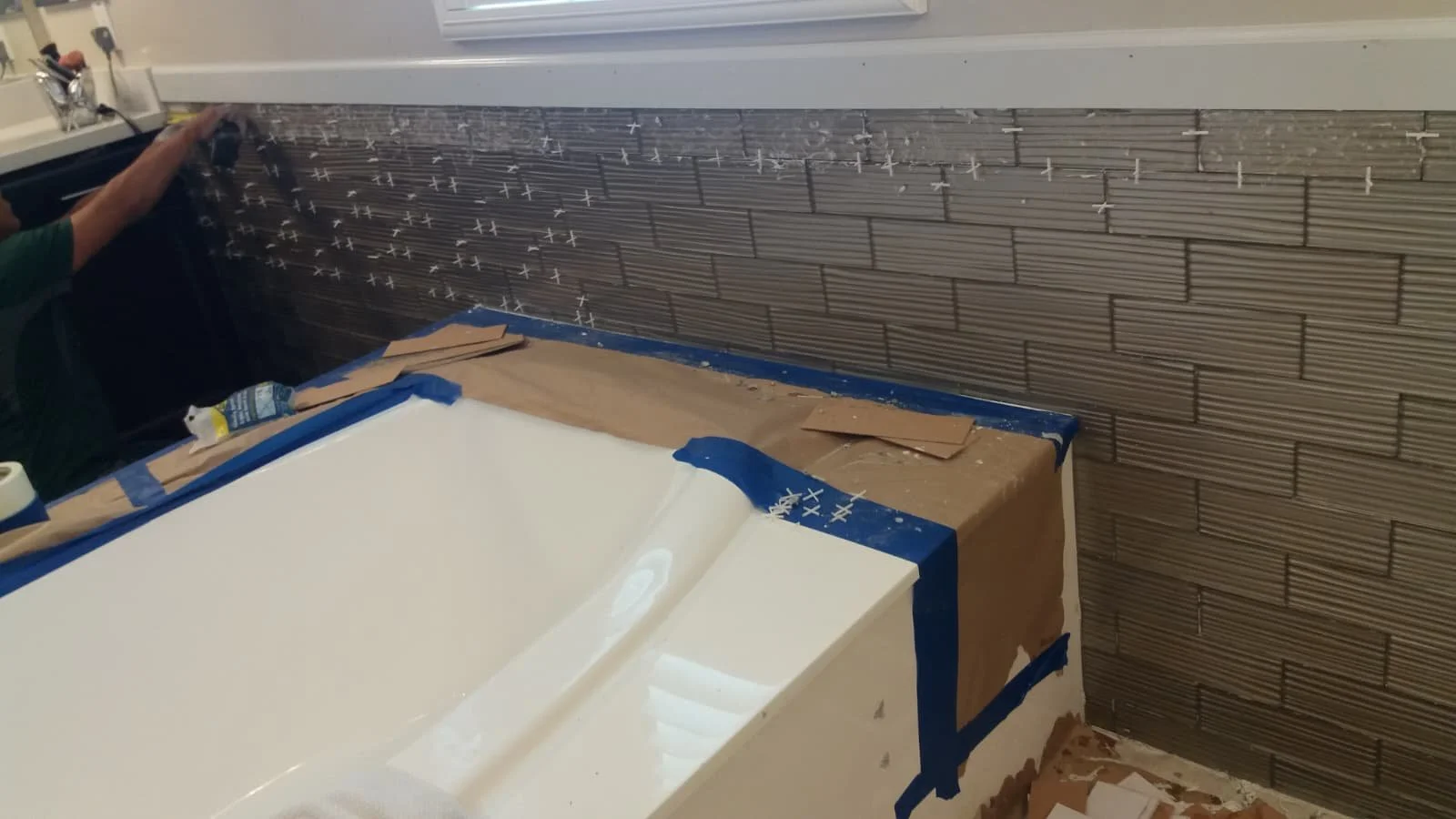 Person installing tile backsplash in a bathroom under a window, with a partially tiled wall and a bathtub below, covered with painter's tape and protective paper.