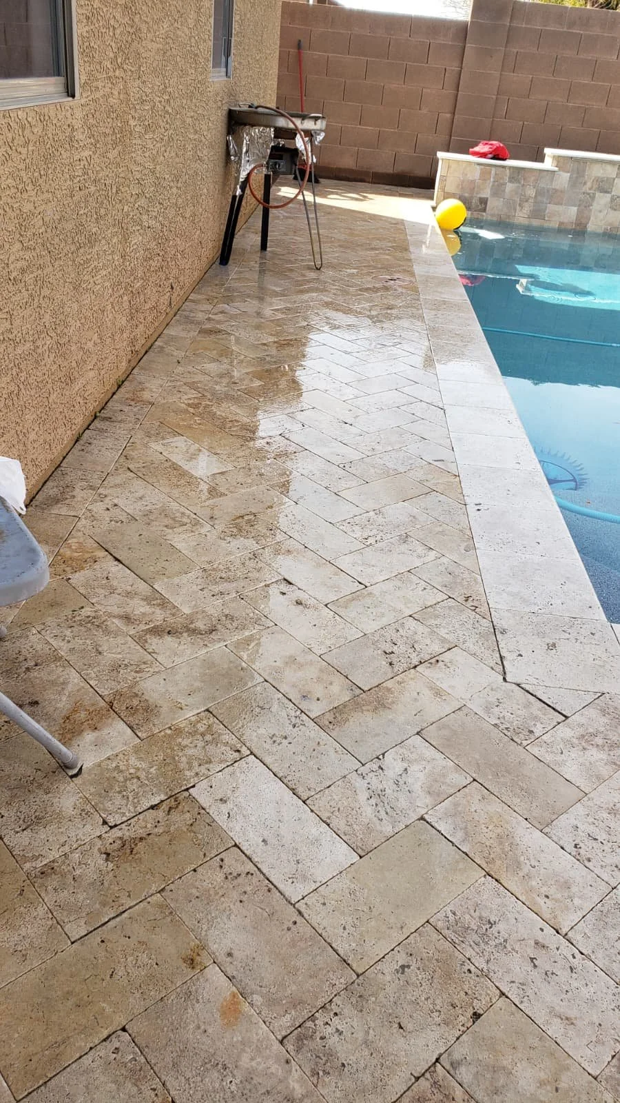 Wet tan tiled poolside patio with a yellow ball and a red cloth on the edge of the pool, a barbecue grill, and a brown brick wall in the background.