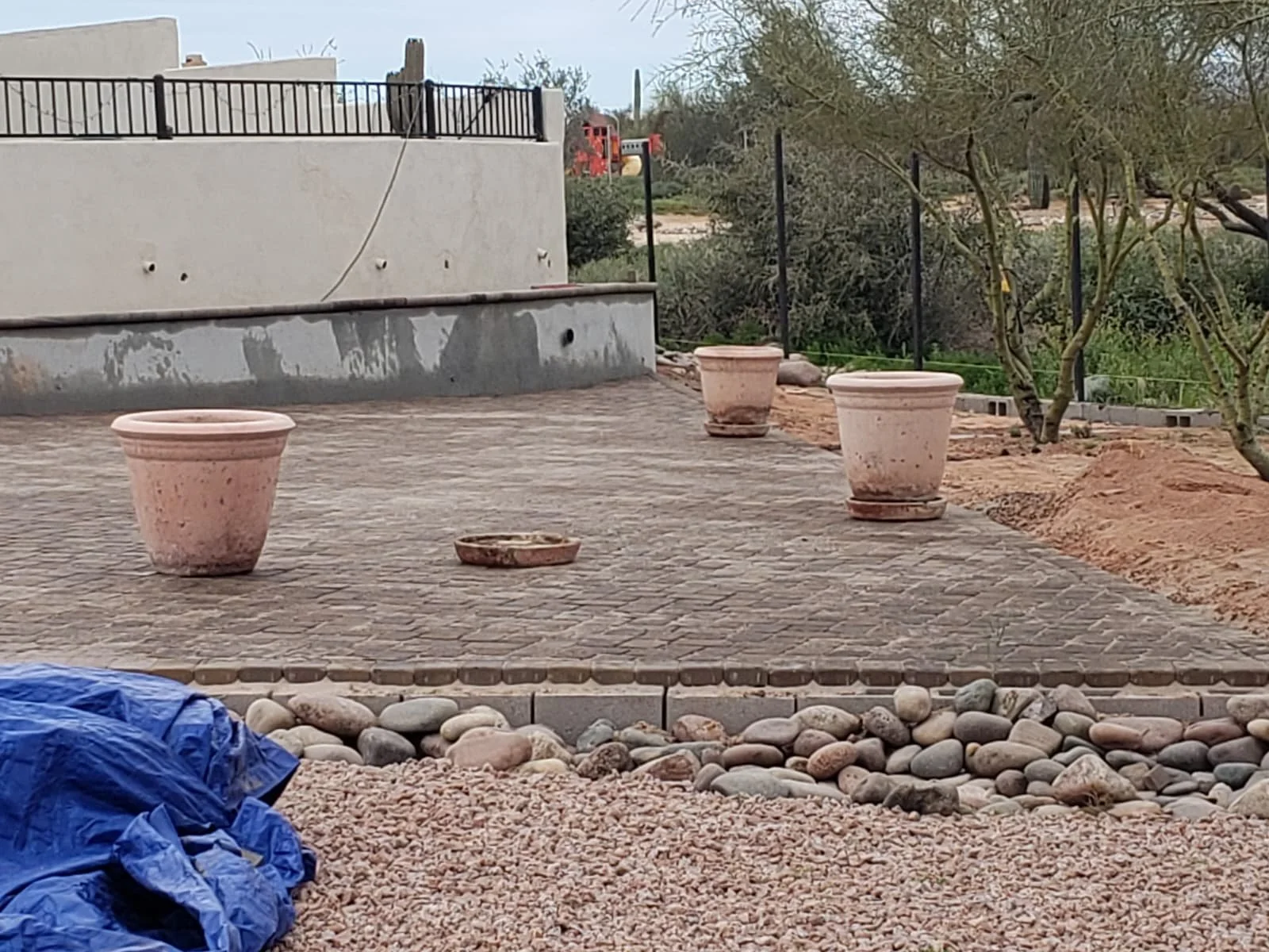 An outdoor patio area with four large empty terracotta planters, one smaller dish, and surrounding rocks, with a house wall and garden in the background.