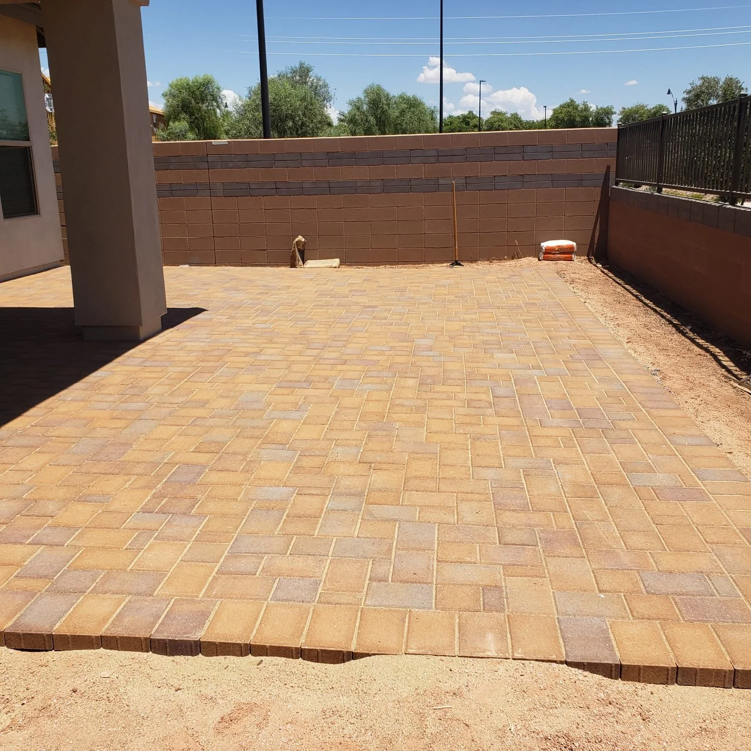 A newly paved brick patio area with some construction tools and materials, surrounded by a brick wall and fencing, under a blue sky with clouds.