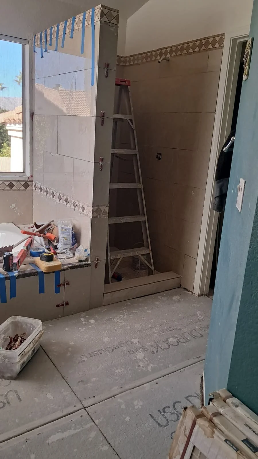 Bathroom under construction with a ladder, tools, and materials, partially tiled wall, window, and a concrete floor with markings.