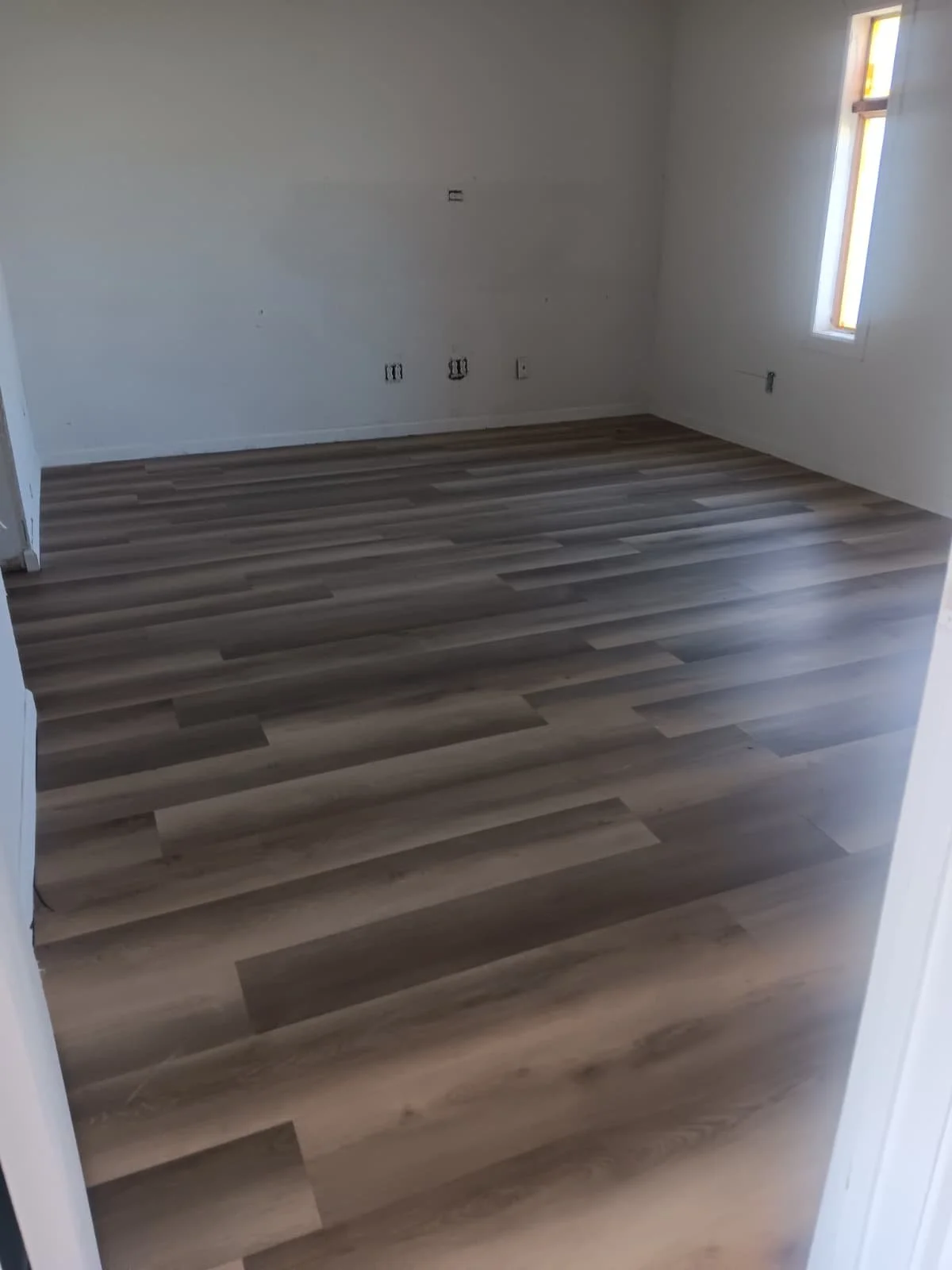 Empty room with light-colored laminate flooring, white walls, and a single window allowing natural light.