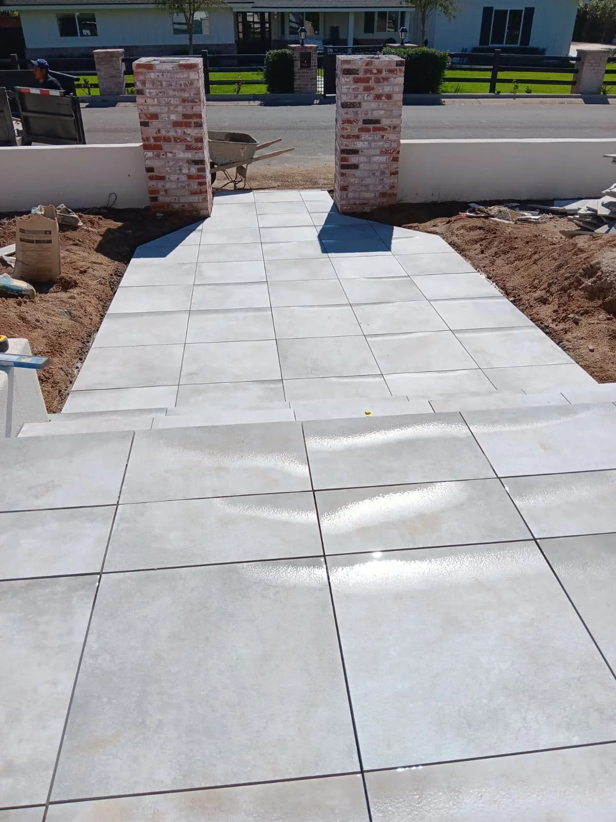 Newly installed white tiled pathway leading to a brick entrance, surrounded by construction materials and dirt, with a street and house in the background.