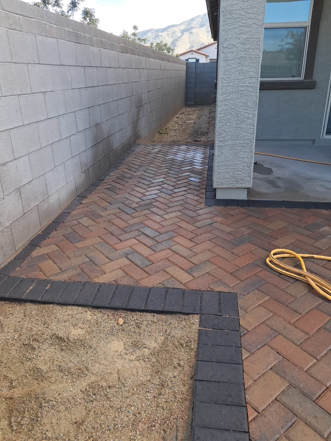 Finished brick walkway next to a house with a concrete wall on one side, a gray interior wall, and mountains in the background.
