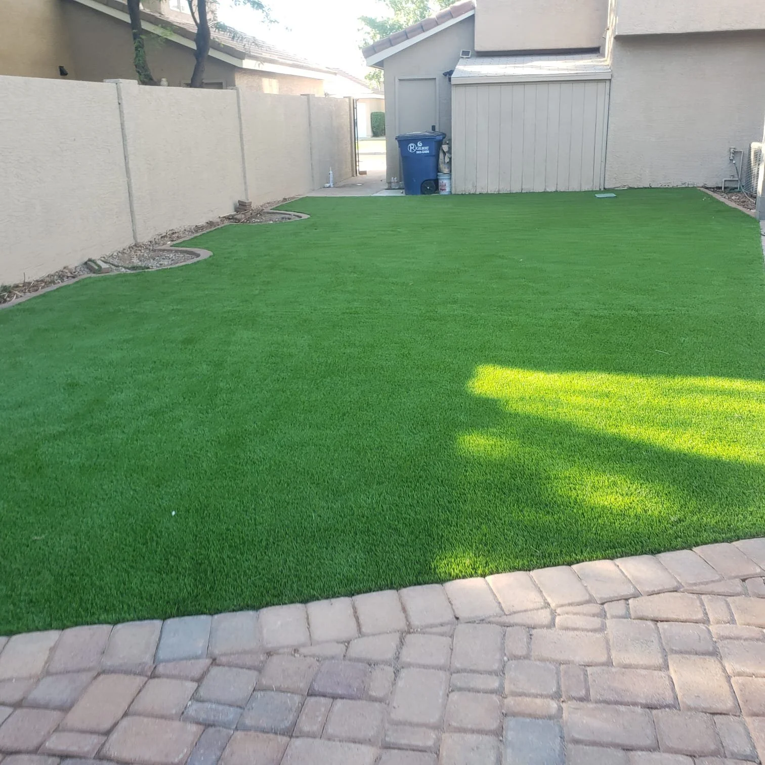 Backyard with a well-maintained green artificial grass lawn, a paved brick patio at the foreground, and a beige house with a small shed and trash bin in the background.