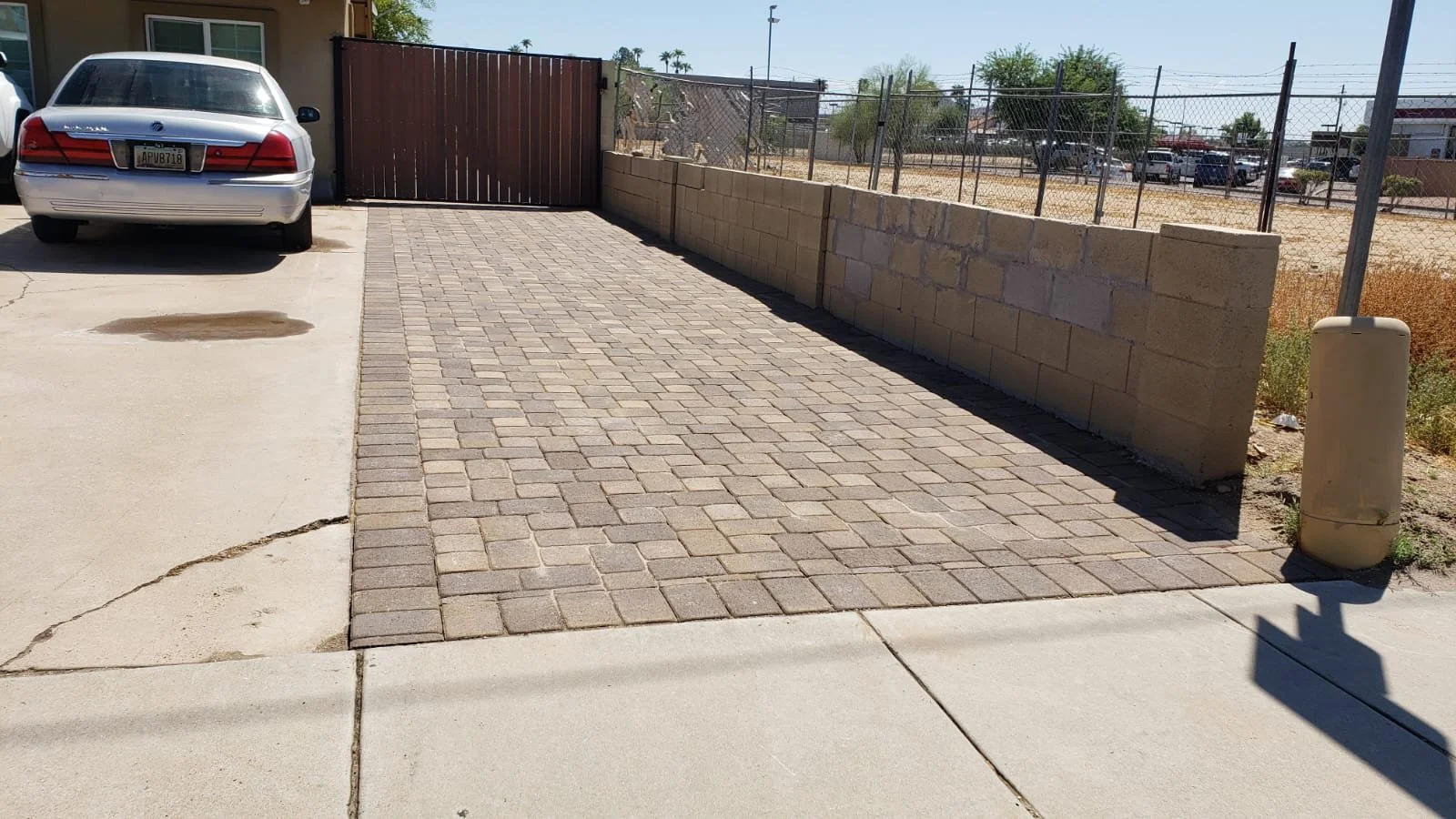 A residential parking area with a silver car parked on the left, a paved walkway with brick pavers, a low beige wall, a chain-link fence, and a street with vehicles in the background under a clear sky.