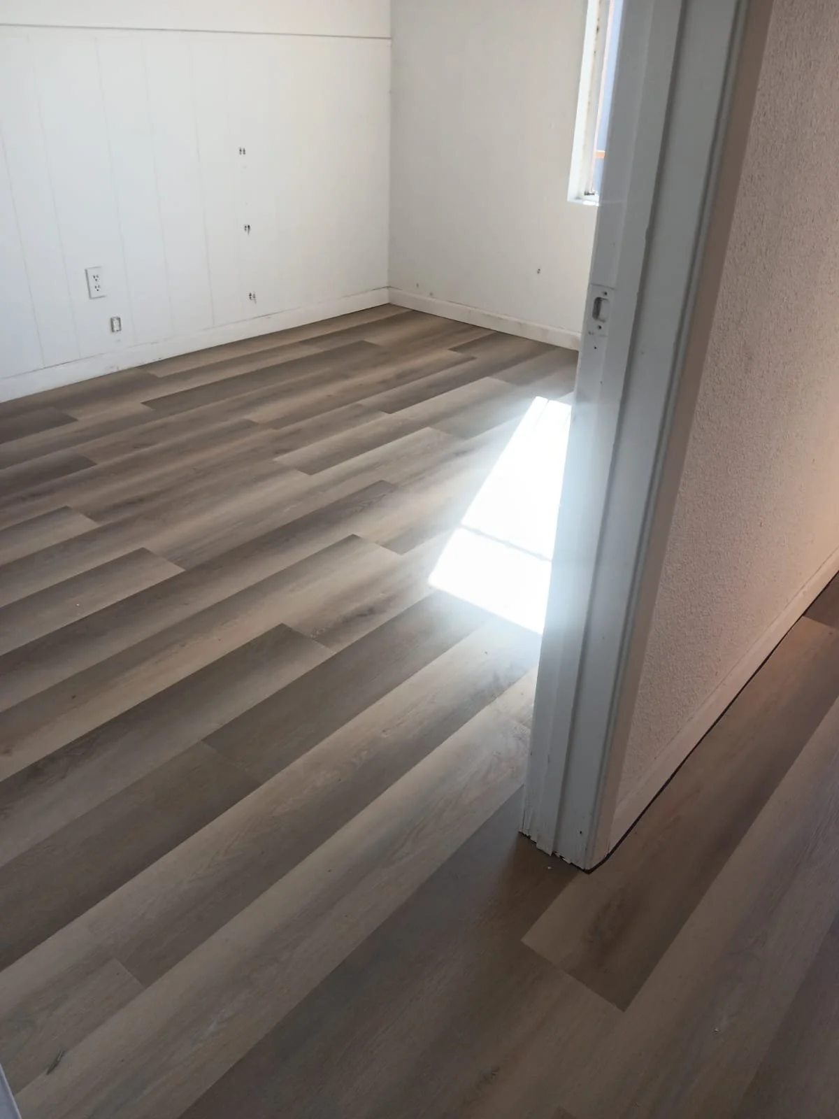 An empty room with light-colored wooden floor, white wall paneling, a window letting in natural light, and electrical outlets on the wall.