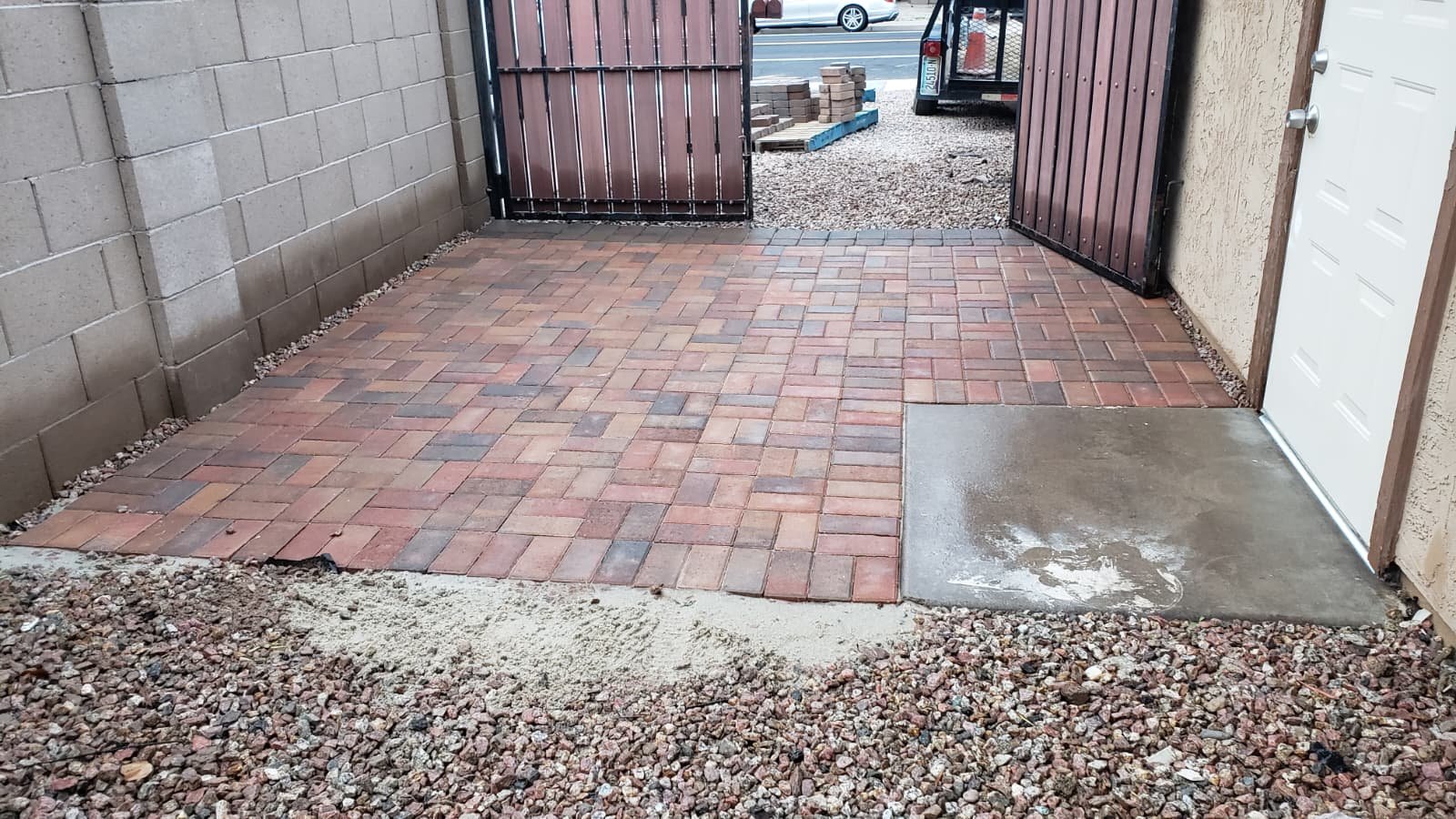 A small entrance with a new brick-paved path leading to a gate, with gravel surrounding the pavement and a concrete pad in front of a white garage door.