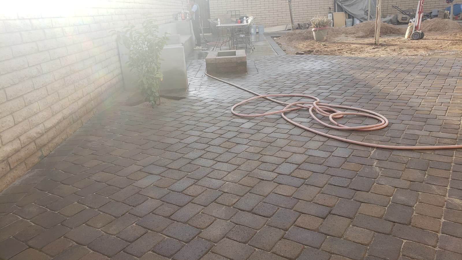 A backyard patio with brick pavers, a garden hose, a table with chairs, and construction materials.