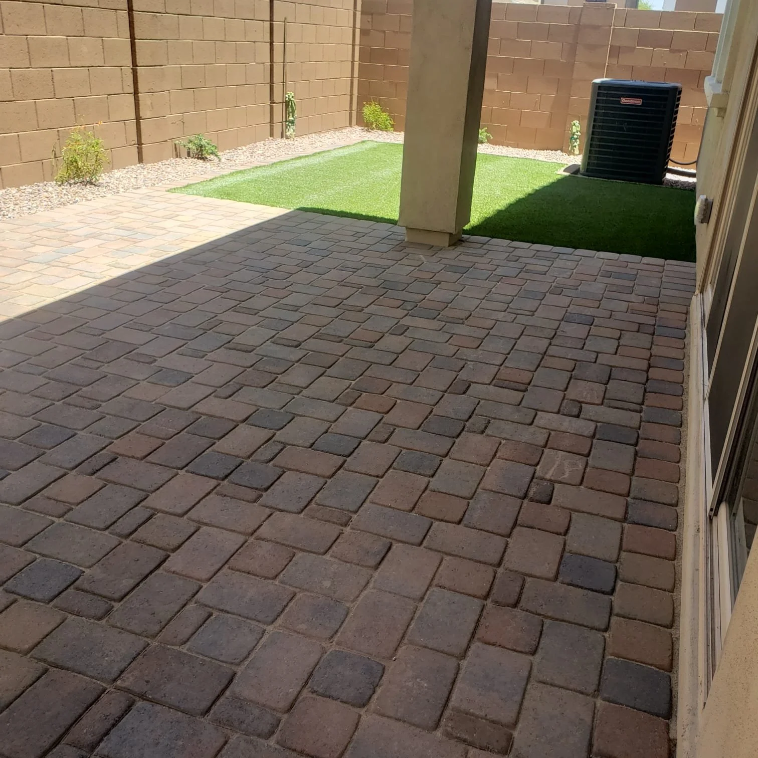 View of a brick patio with a shaded area, a small patch of artificial grass, plants along the wall, and an external air conditioning unit.