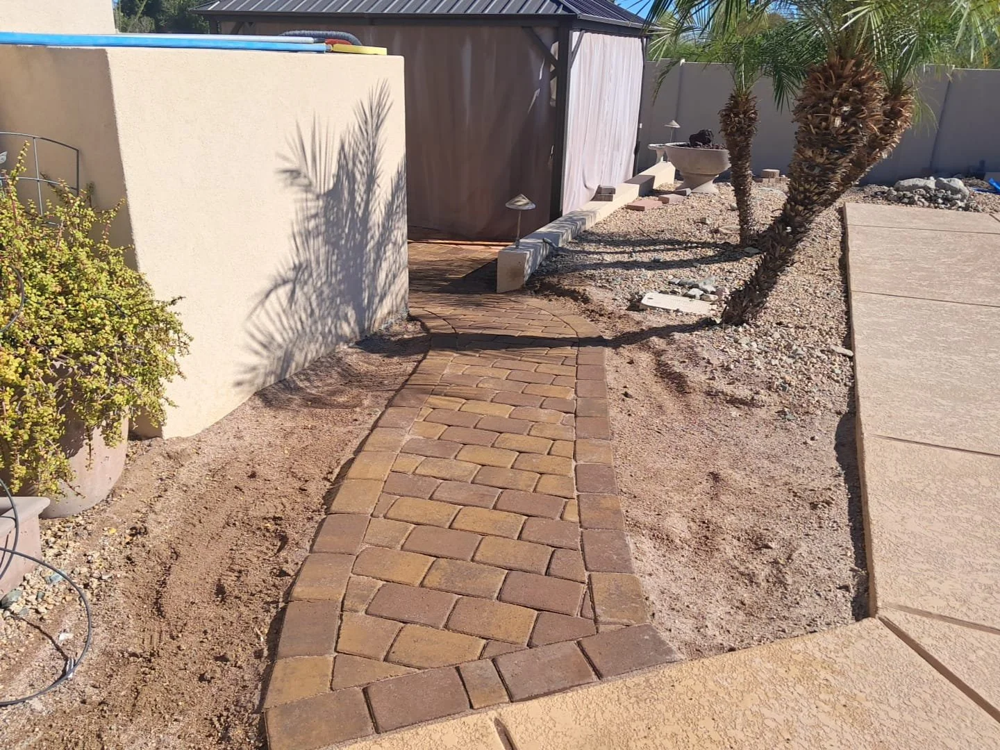 A backyard with a brick walkway, desert plants, and a wooden shed, partially landscaped with gravel and sand.
