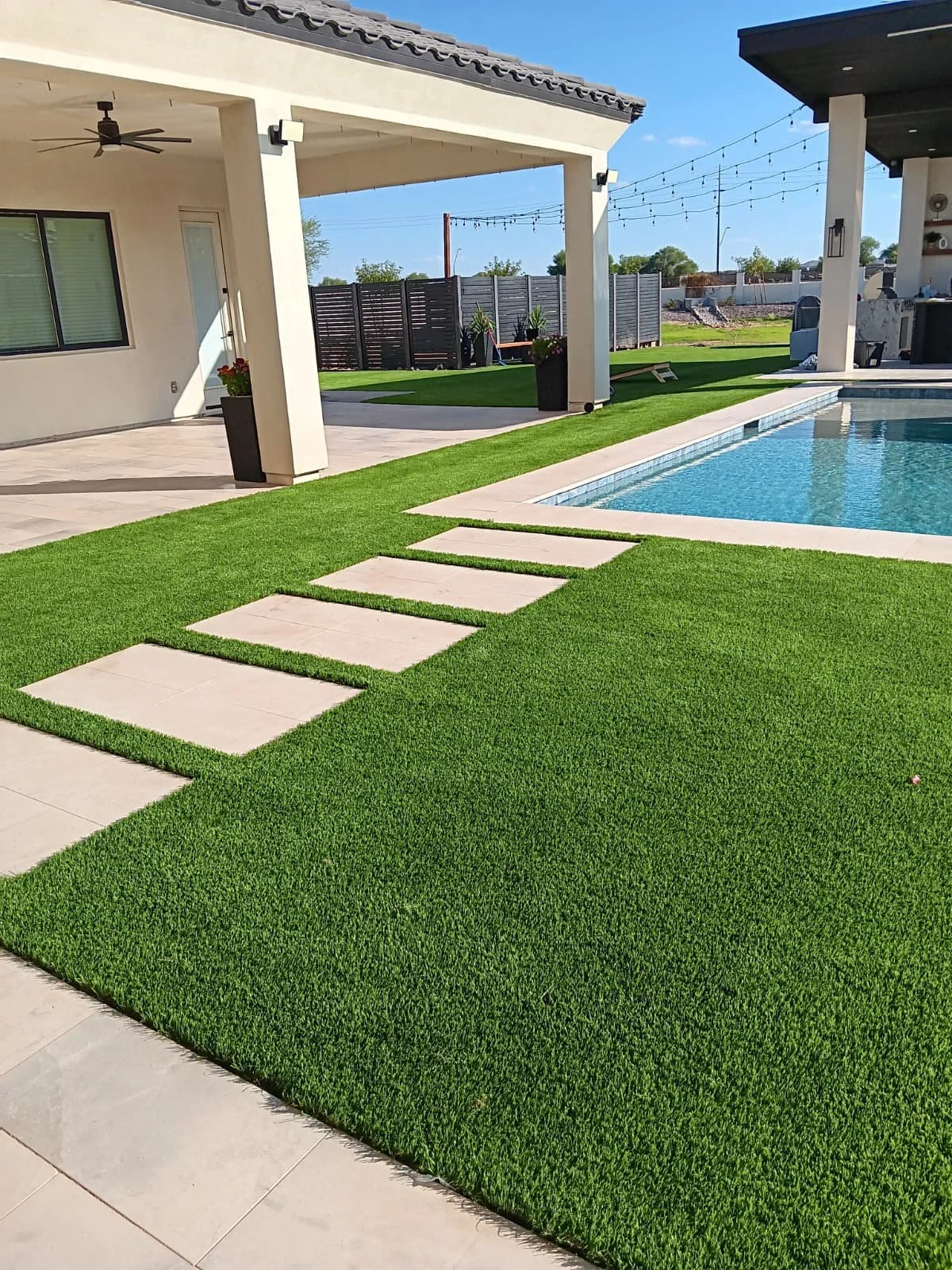 Backyard with a swimming pool, artificial grass, and concrete walkway with stepping stones, covered patio with ceiling fan, and outdoor furniture.