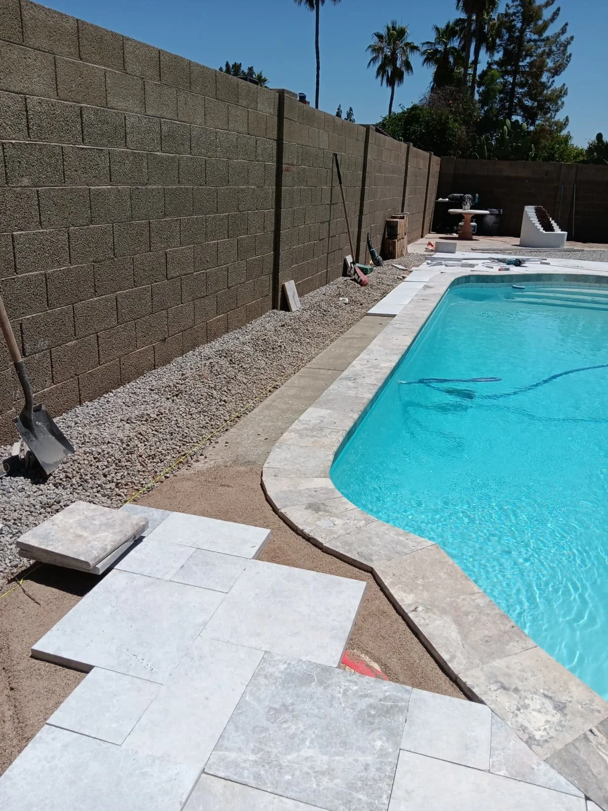 Poolside area with ongoing construction, showing a partially laid stone or tile patio, a blue swimming pool, a brick wall, and various tools and materials.