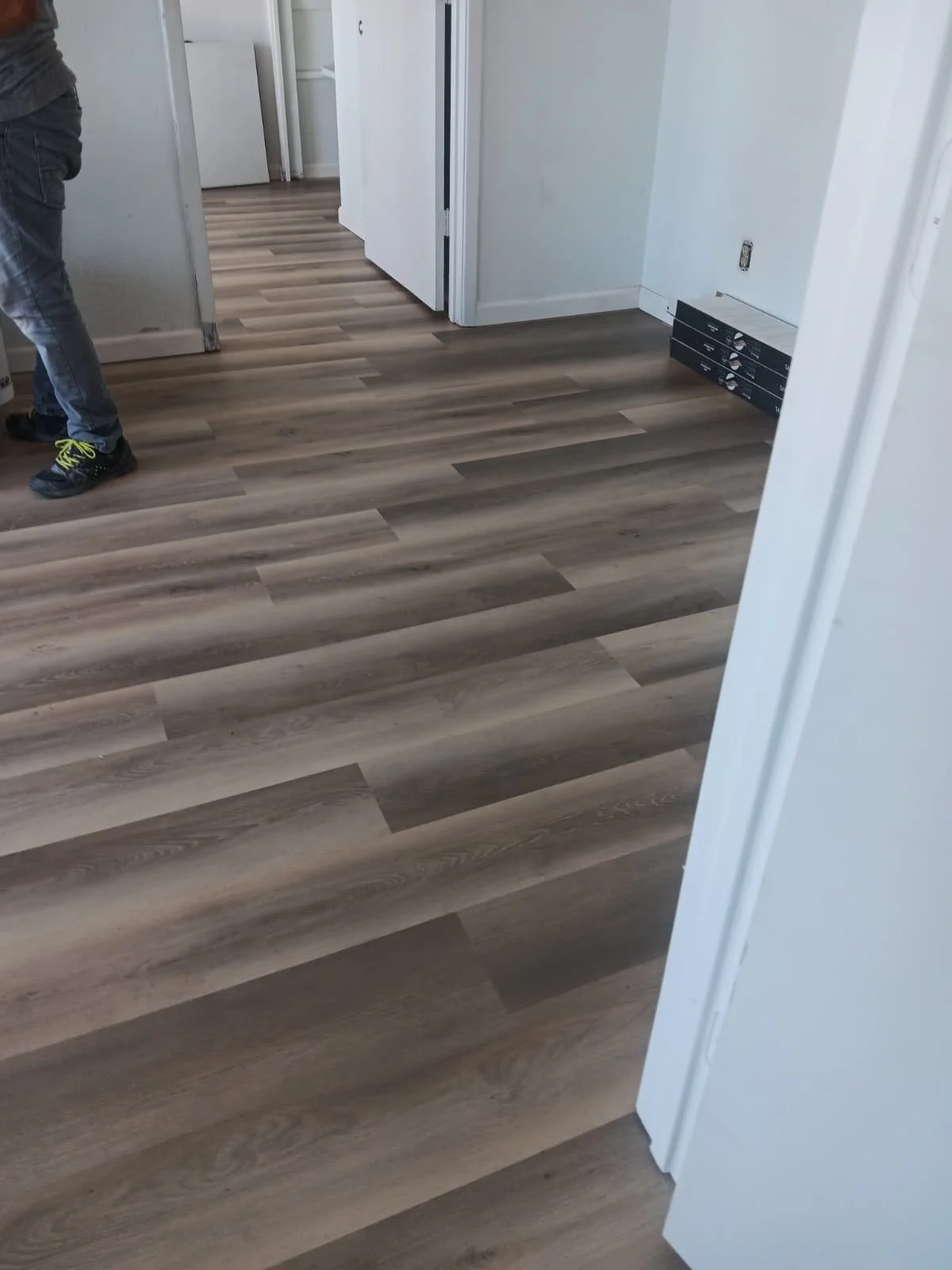 A room with new wood-look laminate flooring, a person standing to the left wearing jeans and black sneakers with neon yellow laces, and a partially open door leading to another room. There's a small box of flooring planks near the wall with an electr