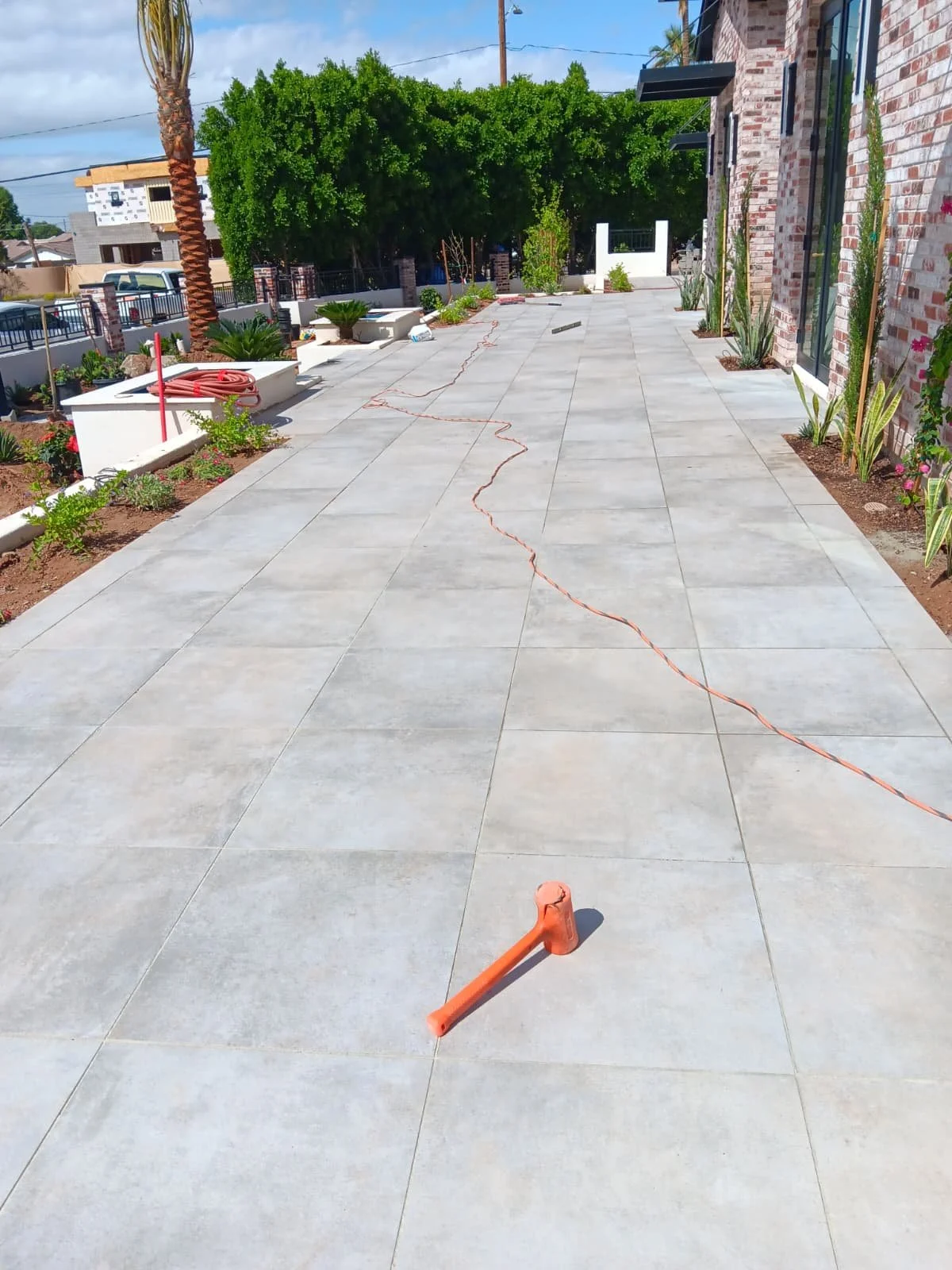 Newly constructed outdoor patio with large gray tiles, surrounded by plants and trees, with a brick wall on the right and construction tools like a hammer and orange cord visible.