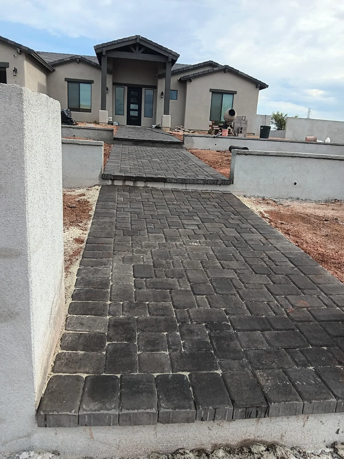 A newly paved stone pathway leading up to a modern house with a covered porch, construction materials and equipment nearby, and cloudy sky overhead.
