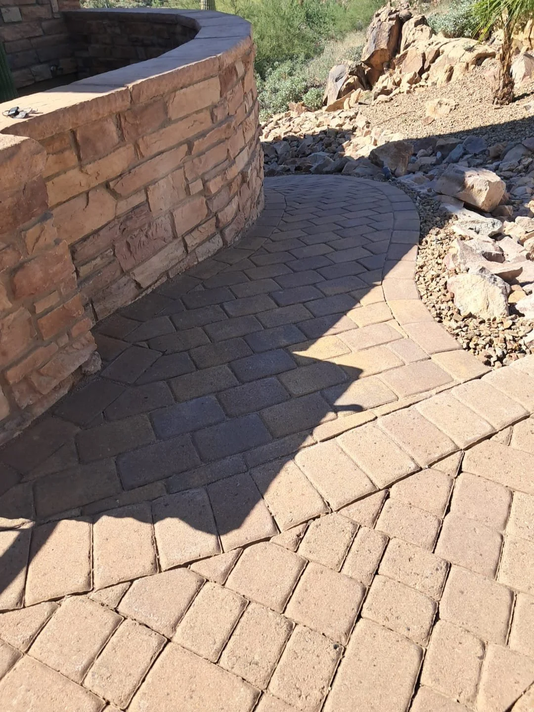 A curved brick walkway next to a stone wall with desert landscaping, rocks, and sparse vegetation in the background.