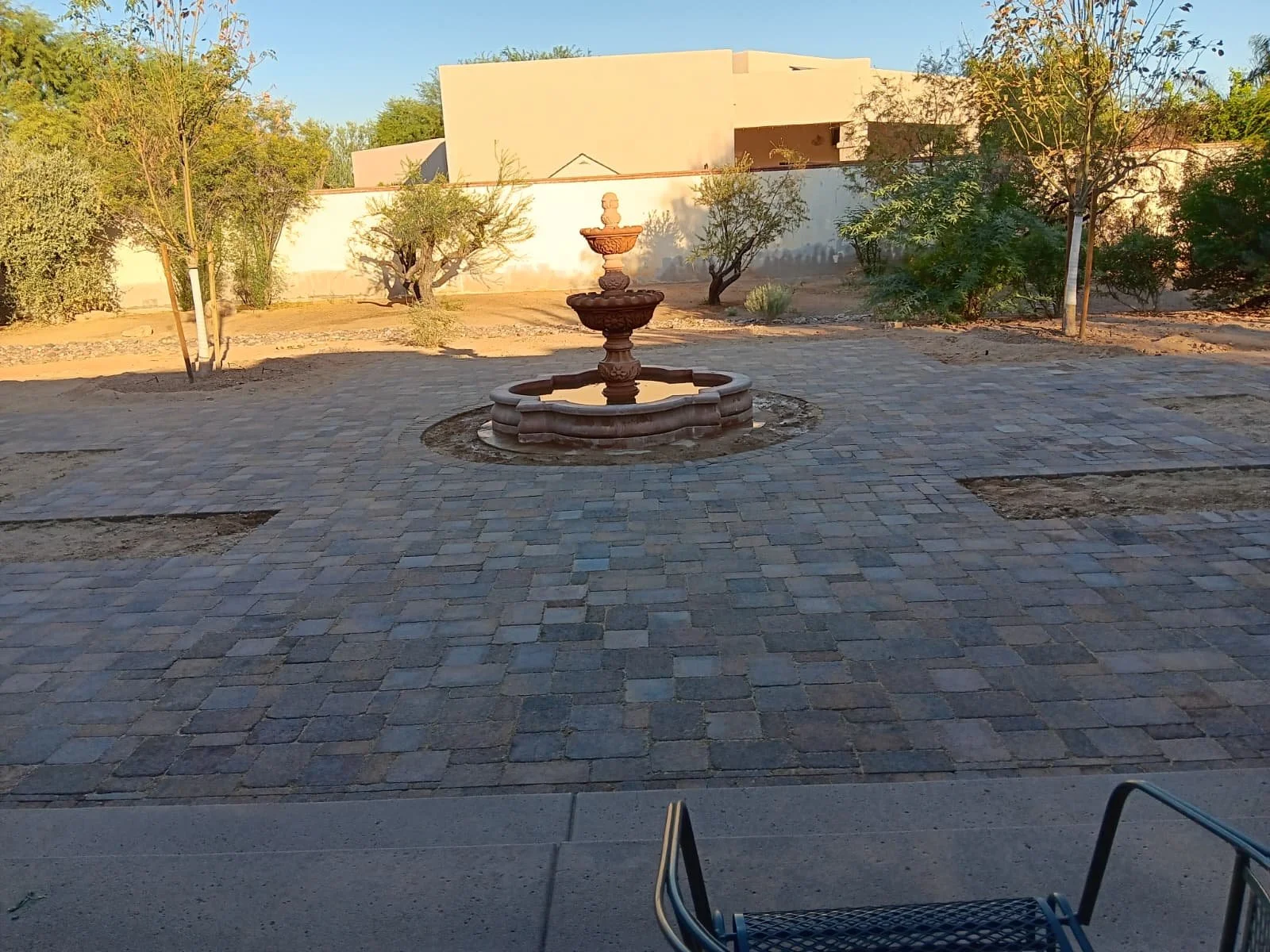 A courtyard with a stone fountain in the center, surrounded by trees and a paved walkway, scene appears to be in a warm, dry climate.