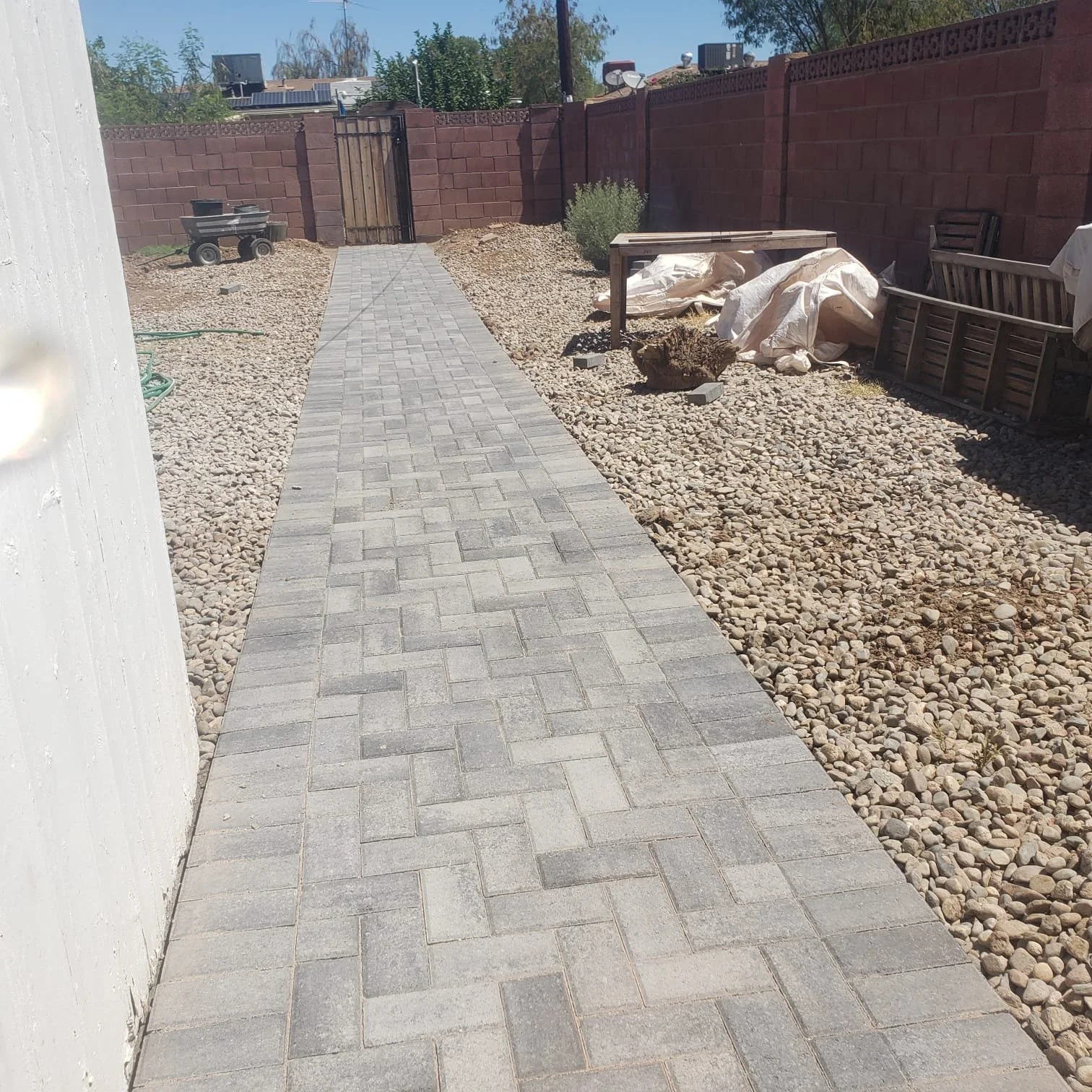 Newly paved brick pathway in a backyard with gravel on both sides, bordered by a white wall on the left and a wooden fence on the right. Construction materials and tools are visible, indicating ongoing yard renovation.