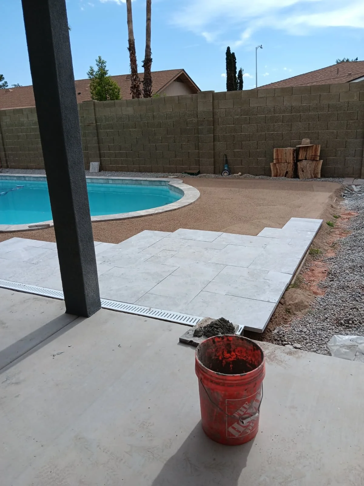 Backyard with a swimming pool, new tile patio under construction, brick wall, and construction materials including a red bucket and some gravel.