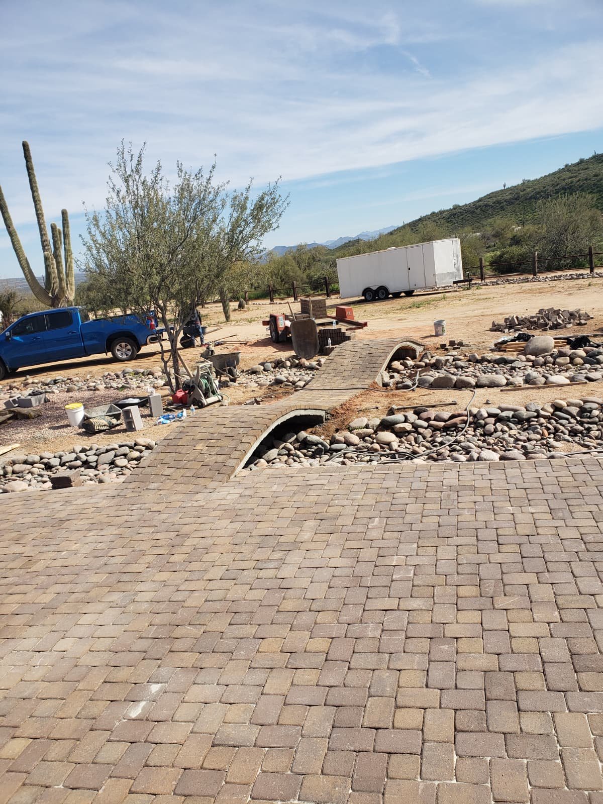 Construction site with a brick walkway, a small bridge over a dry creek bed filled with rocks, a tree, a cactus, and construction equipment, in a desert landscape with mountains in the background.