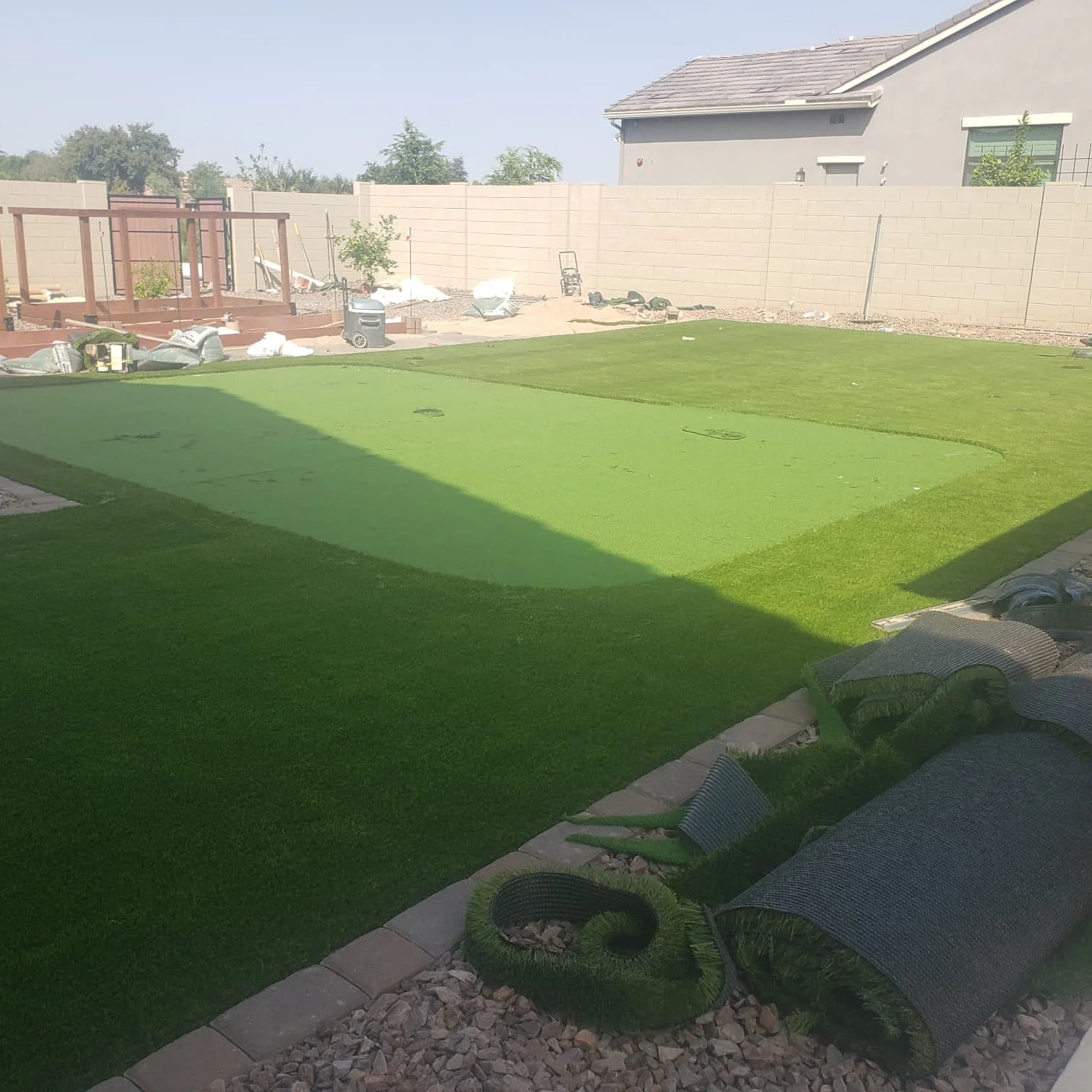 A backyard with a freshly installed green artificial turf lawn, bordered by some patio stones and a gravel area, with construction materials and tools scattered around, and a beige concrete block wall enclosing the yard.
