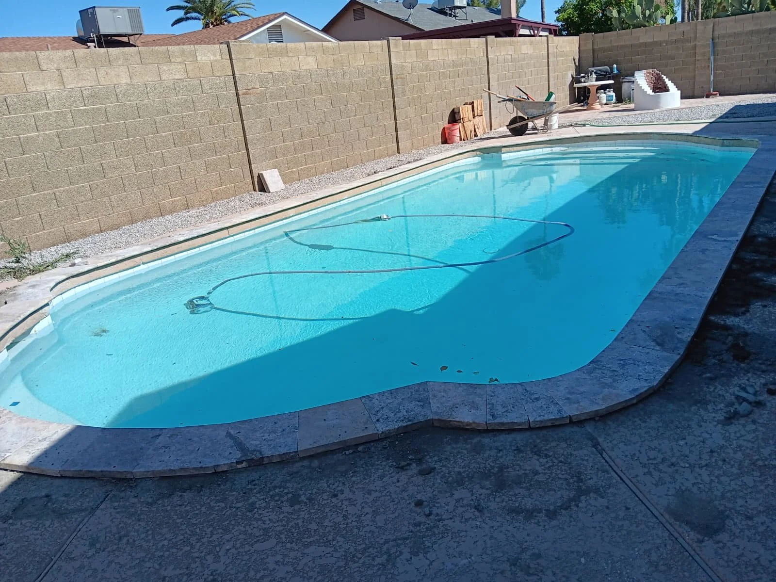 Empty swimming pool in a backyard with a brown brick wall, construction tools and supplies nearby, and a clear blue sky.